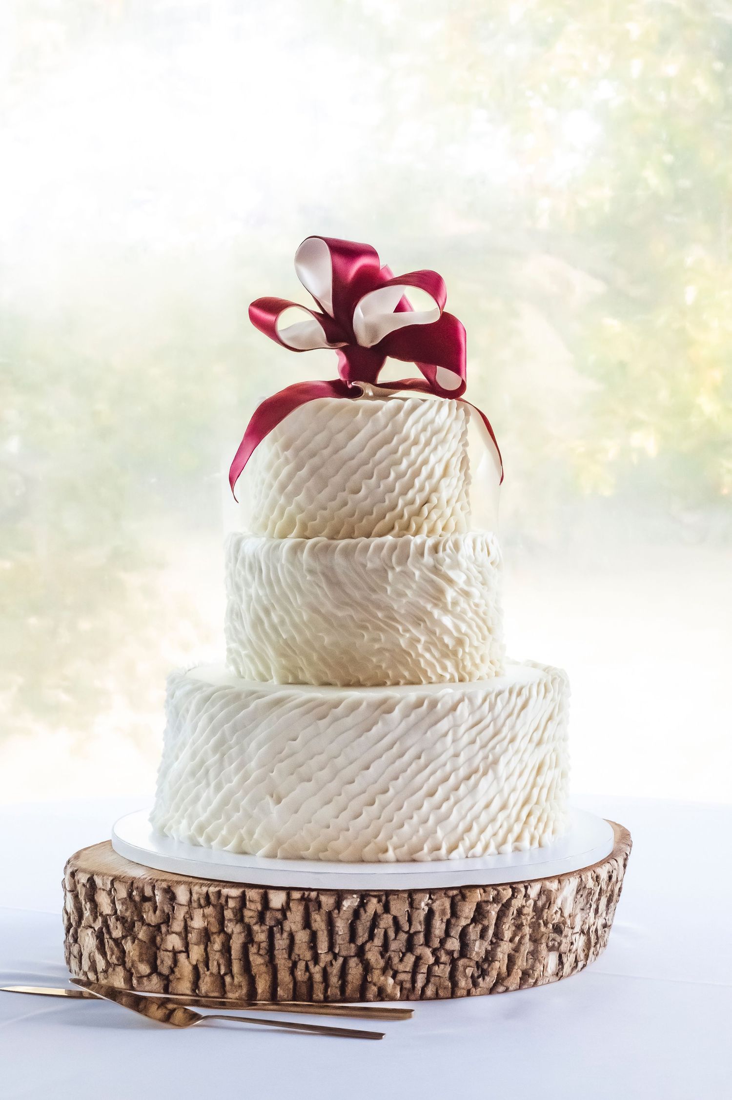 Beautiful three-tier wedding cake topped with a white and red ribbon with a wooden cake stand and gold serving pieces.