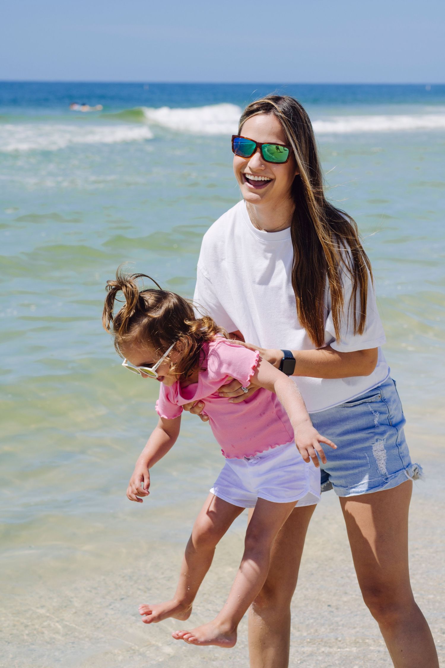 Beachgoers play and laugh together at the ocean's edge on a sunny day with bright blue water and waves.