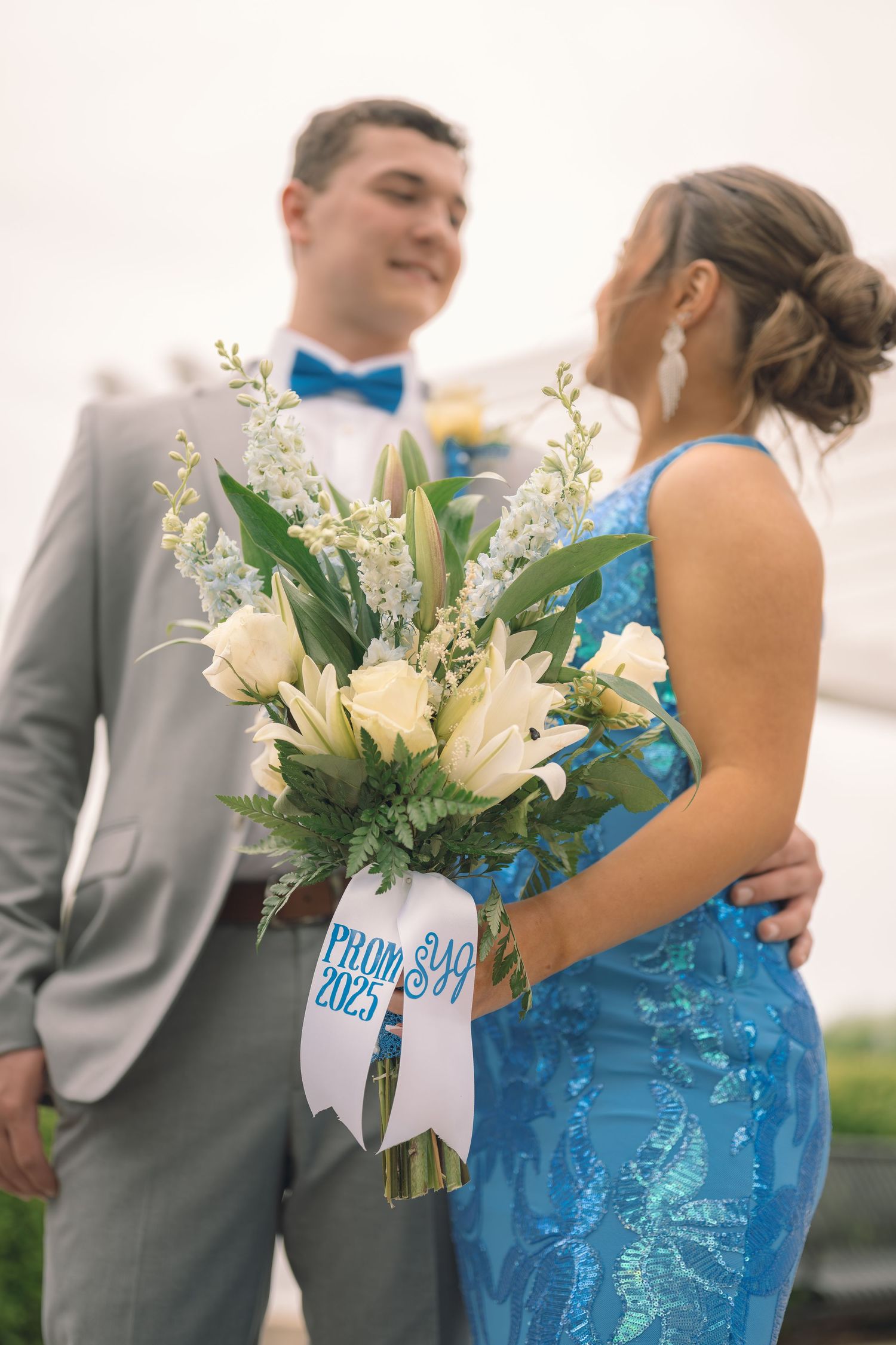 A couple in prom attire embraces outdoors, with the woman holding a white floral bouquet with blue accents and ribbon.