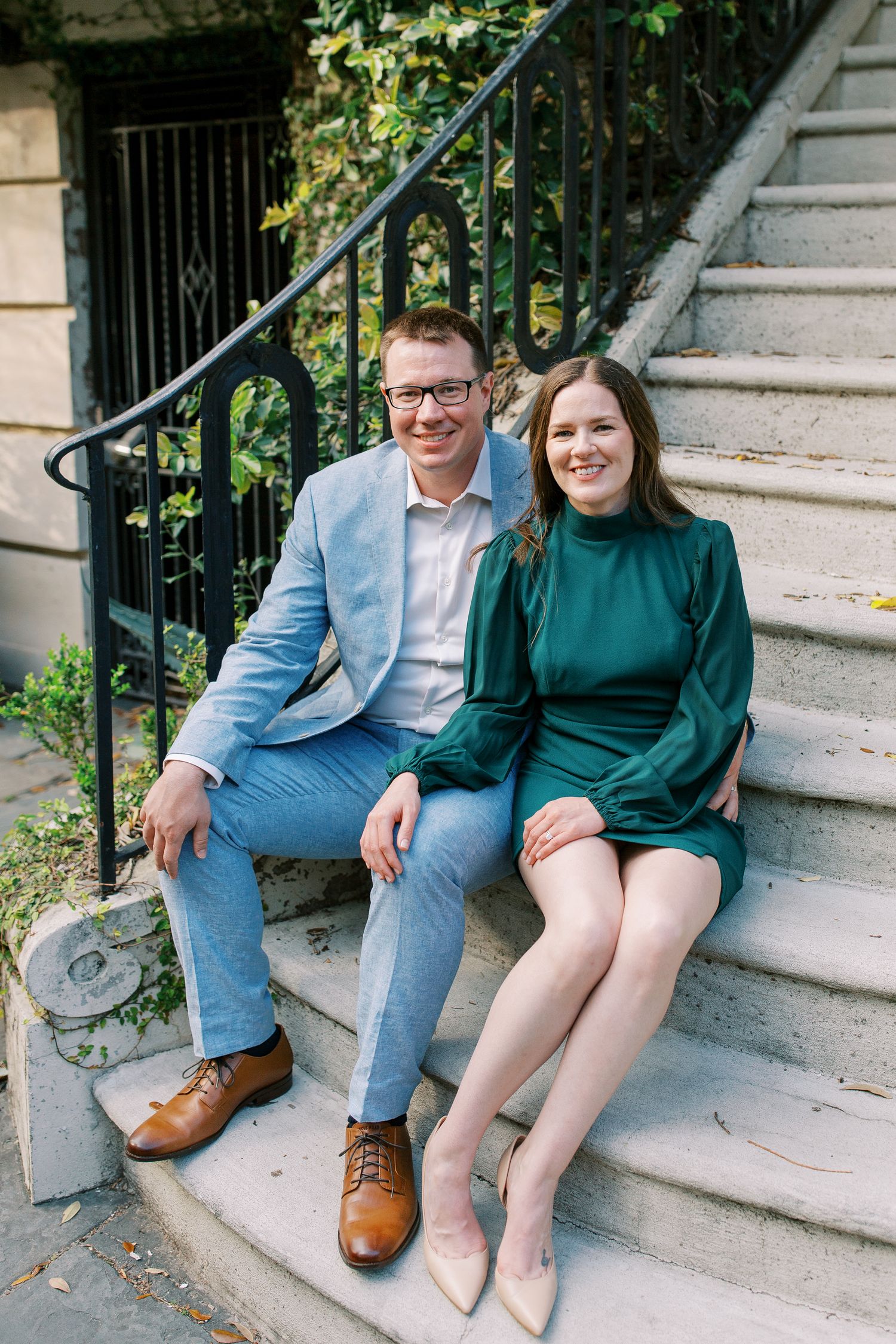A couple in coordinated formal attire sits together on white stone steps with decorative iron railings and vines.