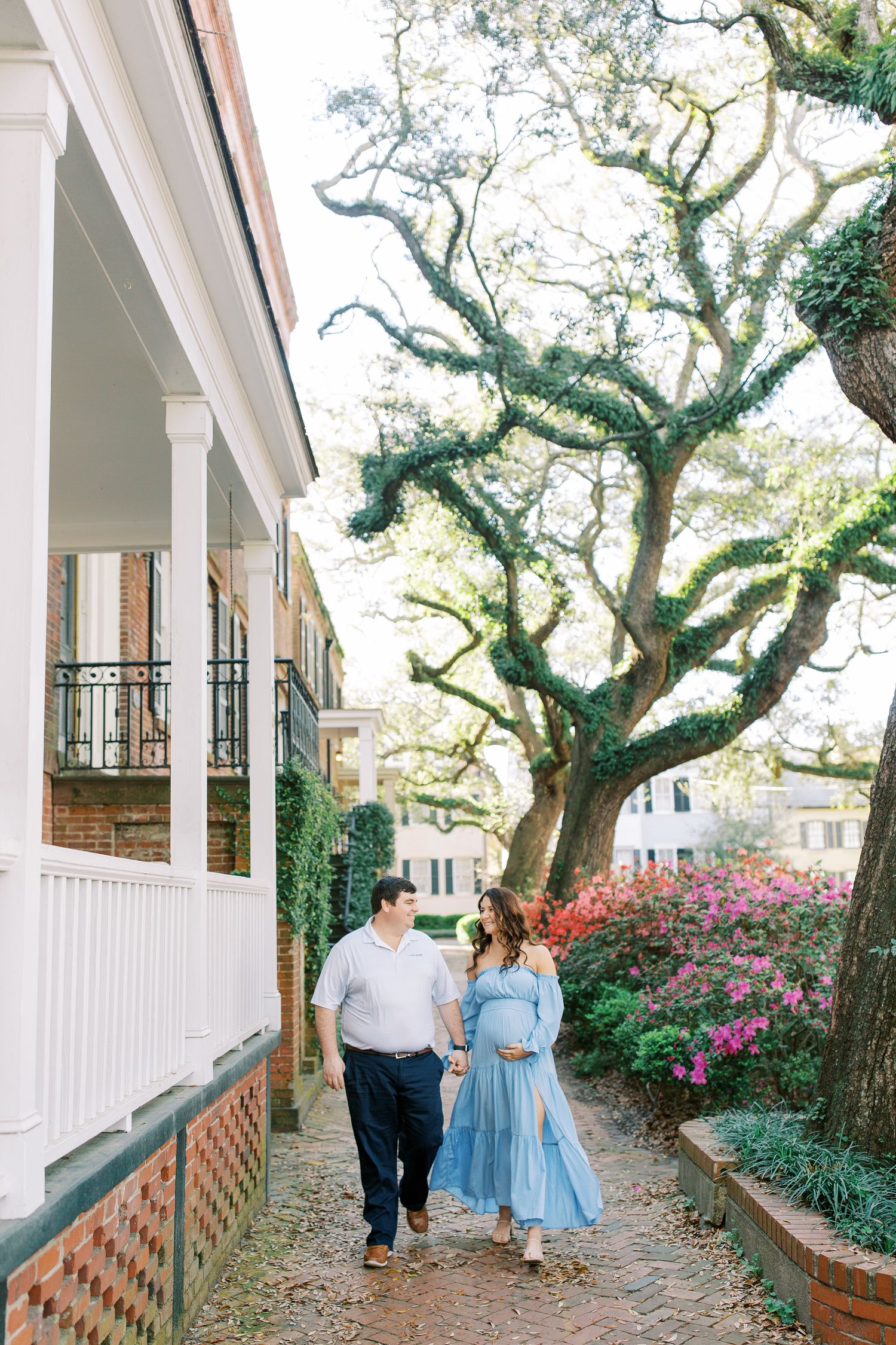 A scenic pathway lined with mossy oak trees and azalea blooms beside a white columned Southern veranda.
