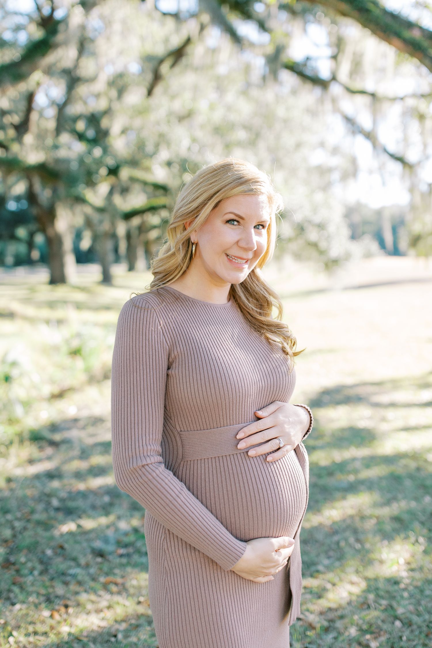 A pregnant woman in a mauve ribbed sweater dress stands outdoors among trees with Spanish moss on an autumn day.
