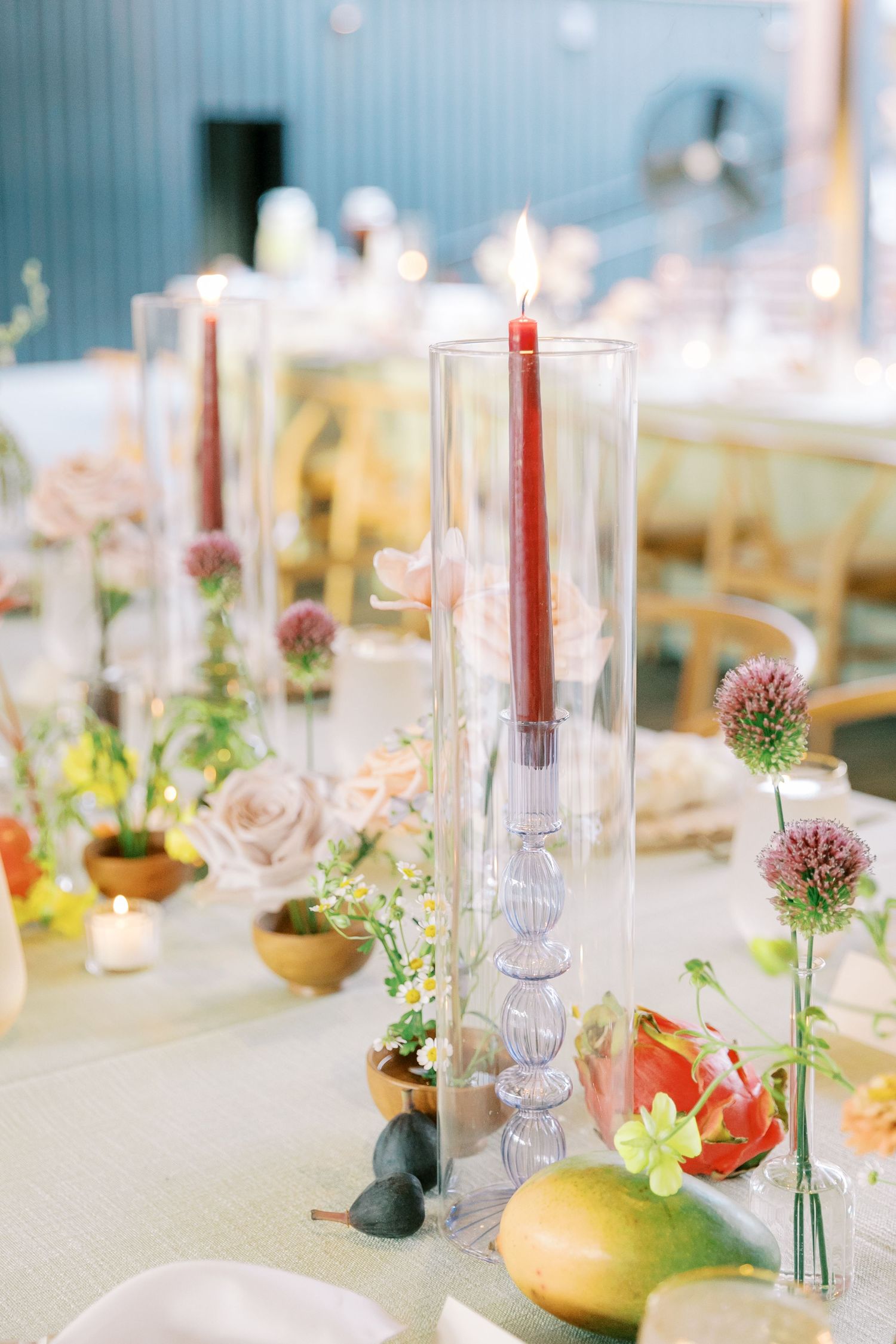 A wedding table setting with red taper candles, fresh flowers, and fruit decorations in a light-filled space.