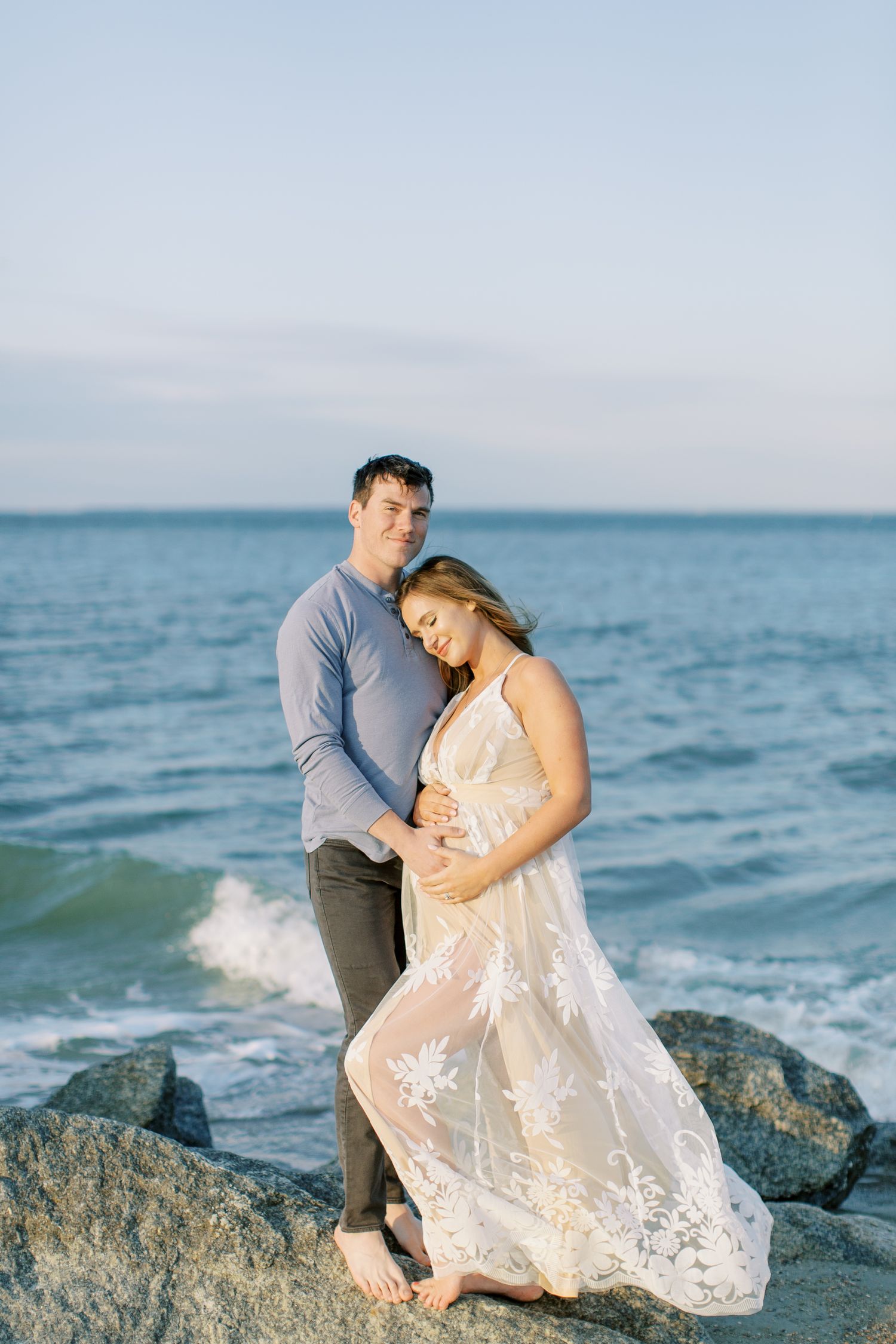 A couple poses romantically on coastal rocks at sunset while gentle waves crash against the shore.