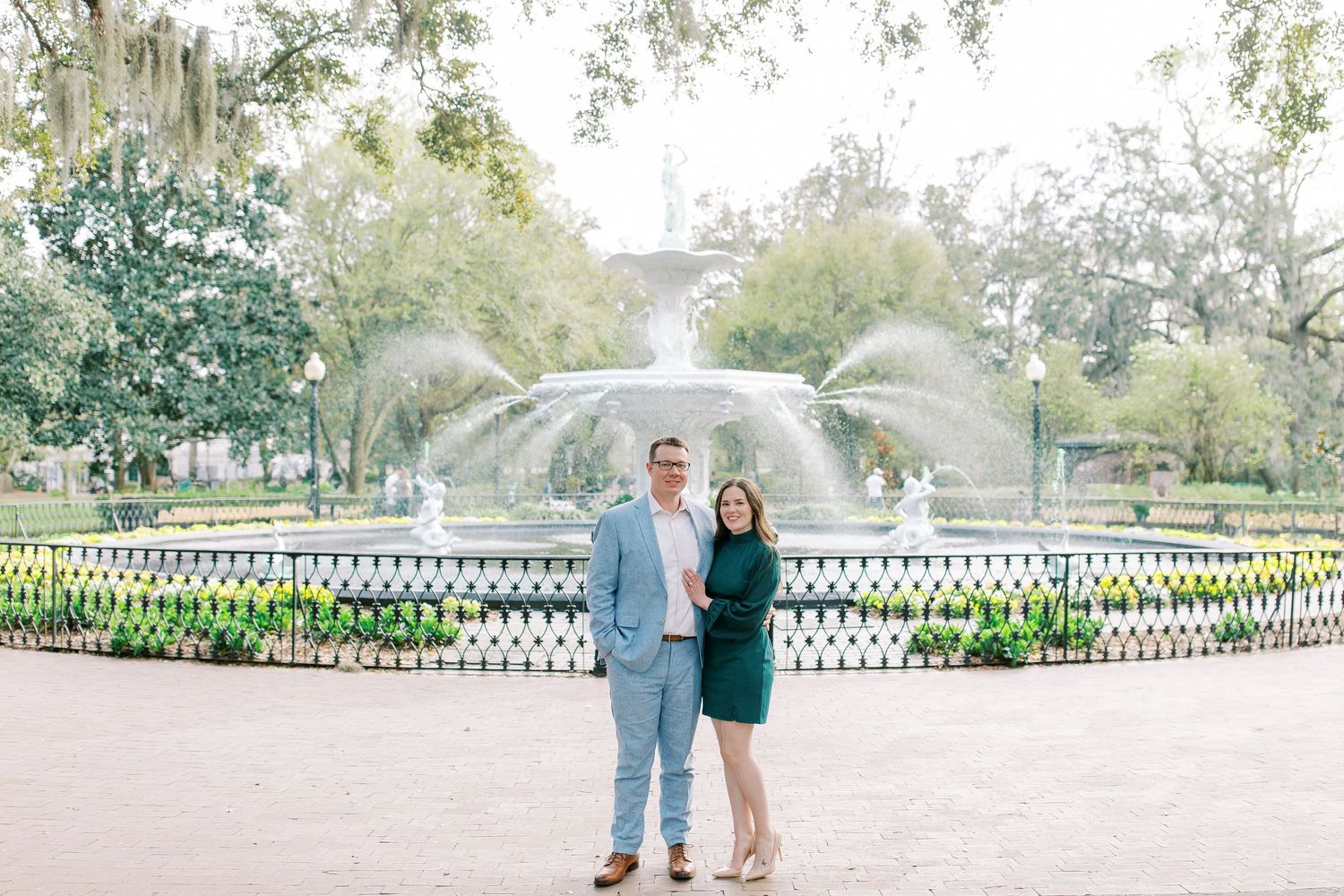 A couple poses in front of Forsyth Park fountain surrounded by gardens and floral landscaping in Savannah, Georgia.