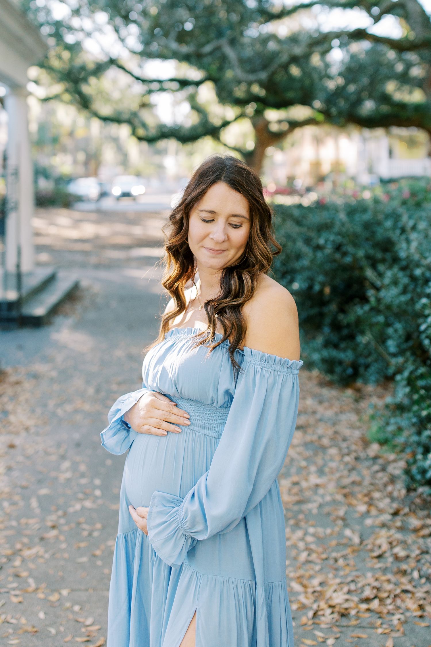 A woman in a blue maternity dress cradles her belly while standing on a leaf-covered path.