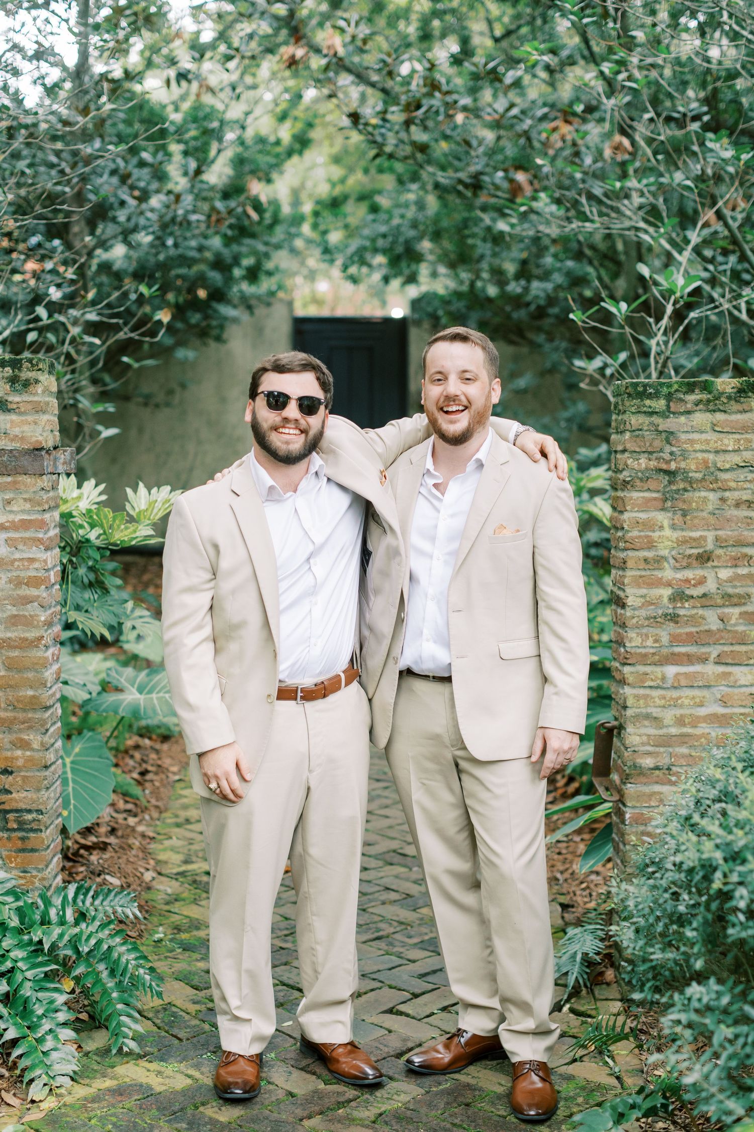 Two friends in matching beige suits pose together in a garden setting with brick walls and greenery.