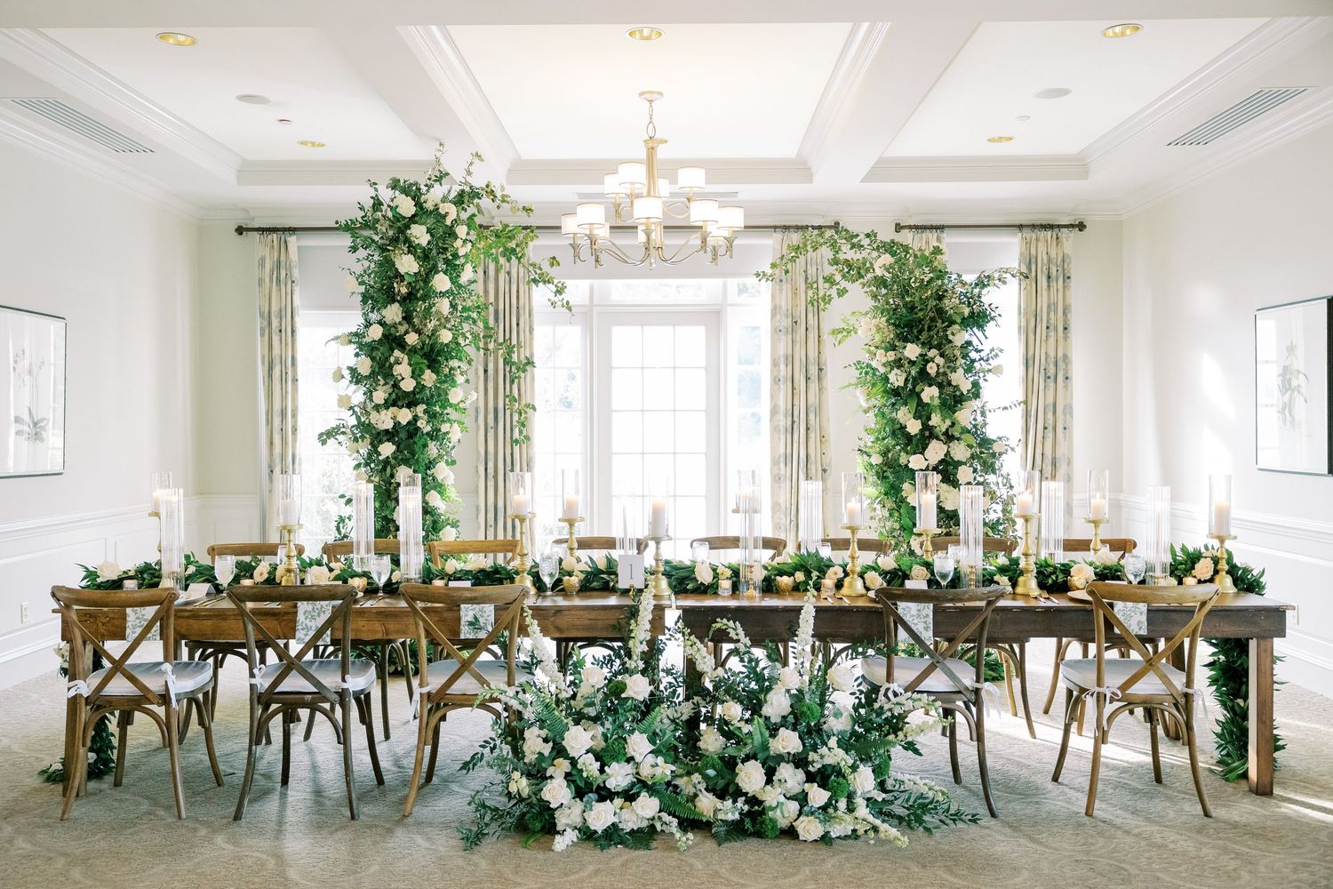 Elegant wedding reception table with lush white floral arrangements and wooden crossback chairs in a bright white room.