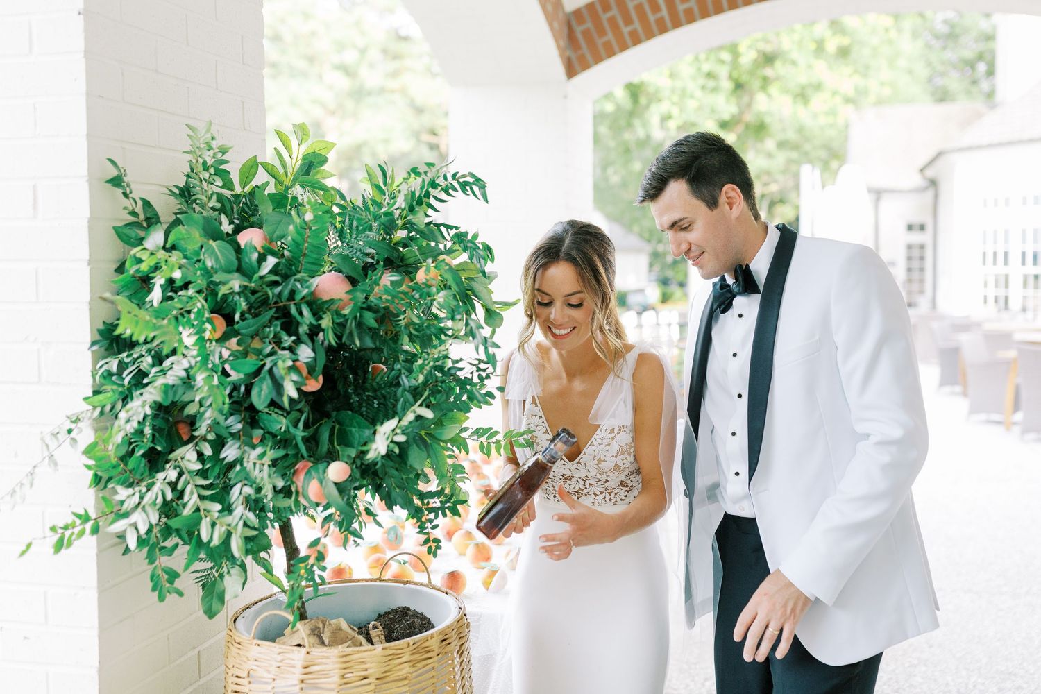 Newlyweds share intimate moment by potted greenery with bride in white gown and groom in white tuxedo jacket.