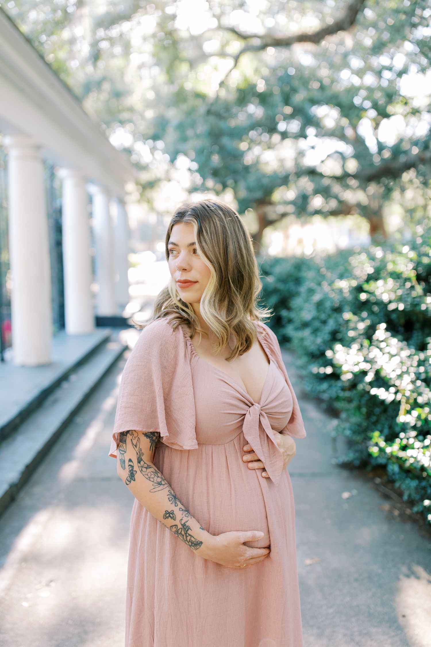 A pregnant woman in a flowing pink dress stands on a sunlit porch with white columns and lush greenery in the background.