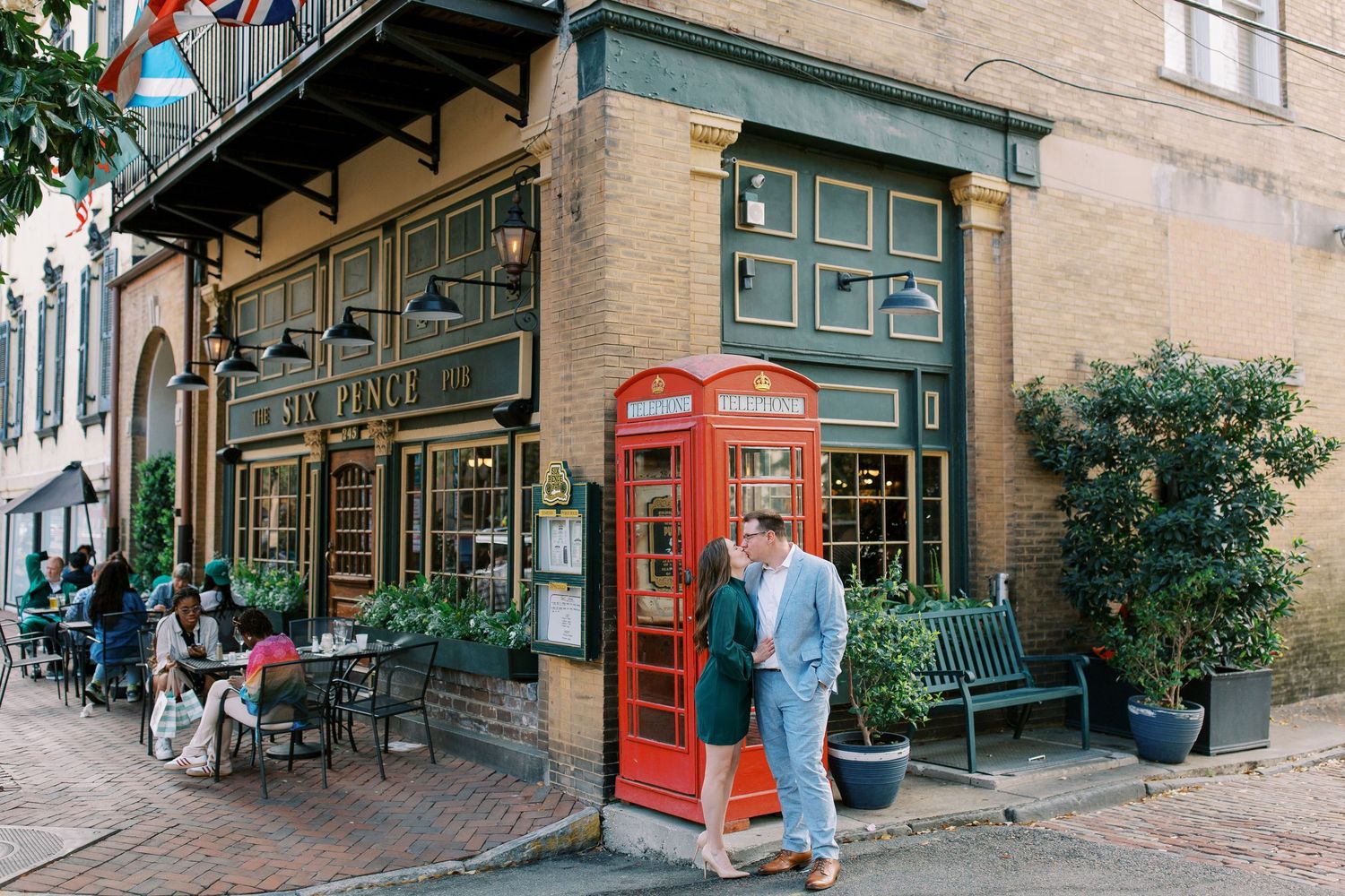 A couple stands near a classic red phone booth outside a charming storefront with green trim and outdoor seating.