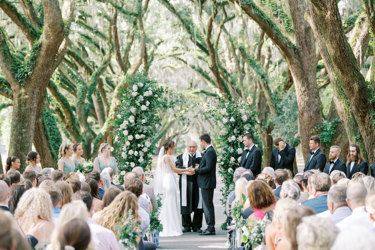Outdoor wedding ceremony under a natural canopy of oak trees decorated with floral arrangements.