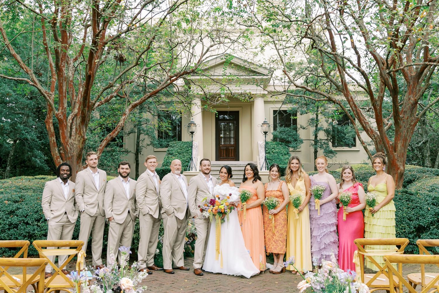 A wedding party in khaki suits and colorful dresses poses on a wooden bridge in front of a classical stone building.