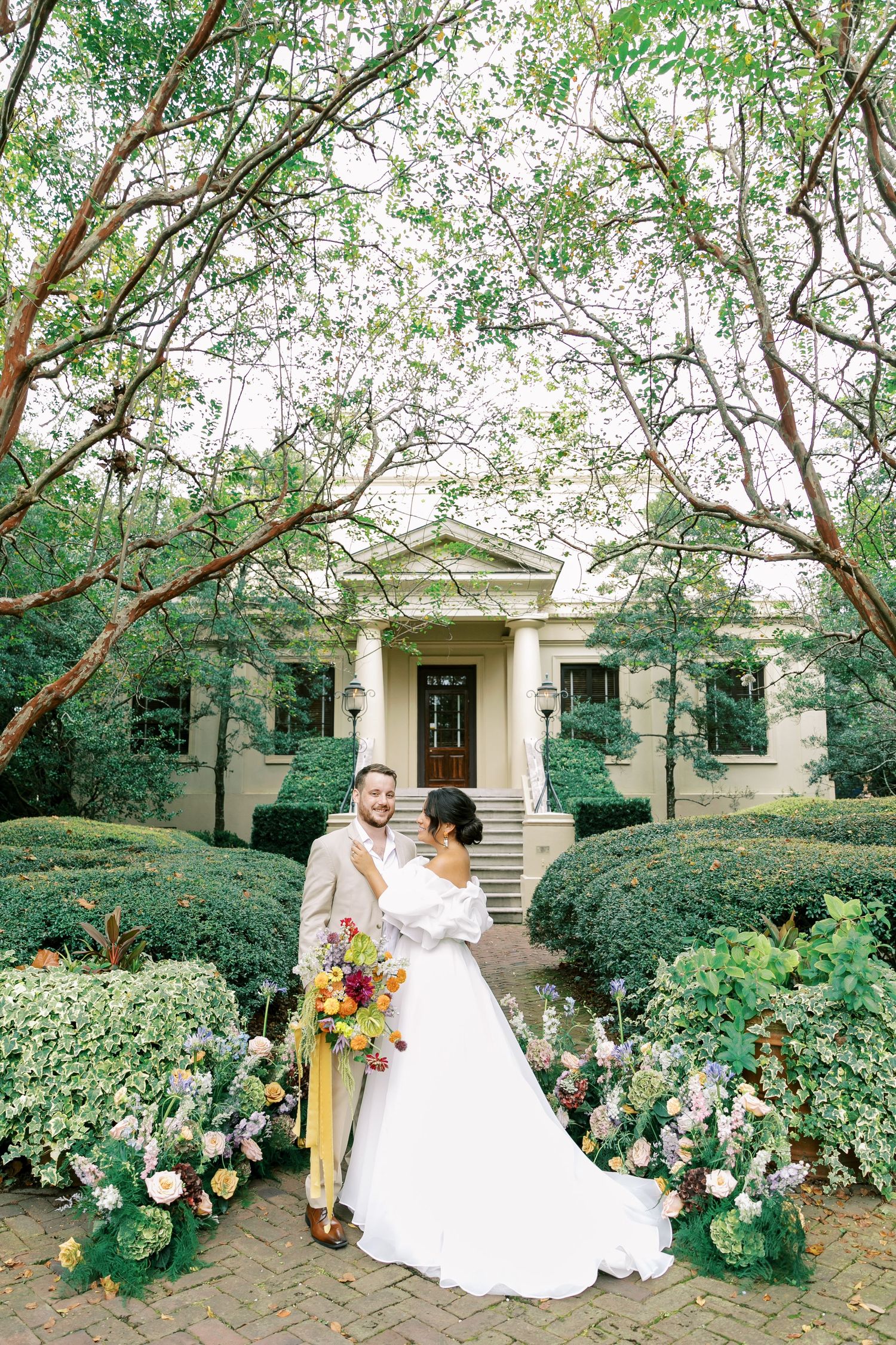 The newlyweds pose in front of an elegant colonial-style mansion with a beautiful garden path lined with blooming flowers.