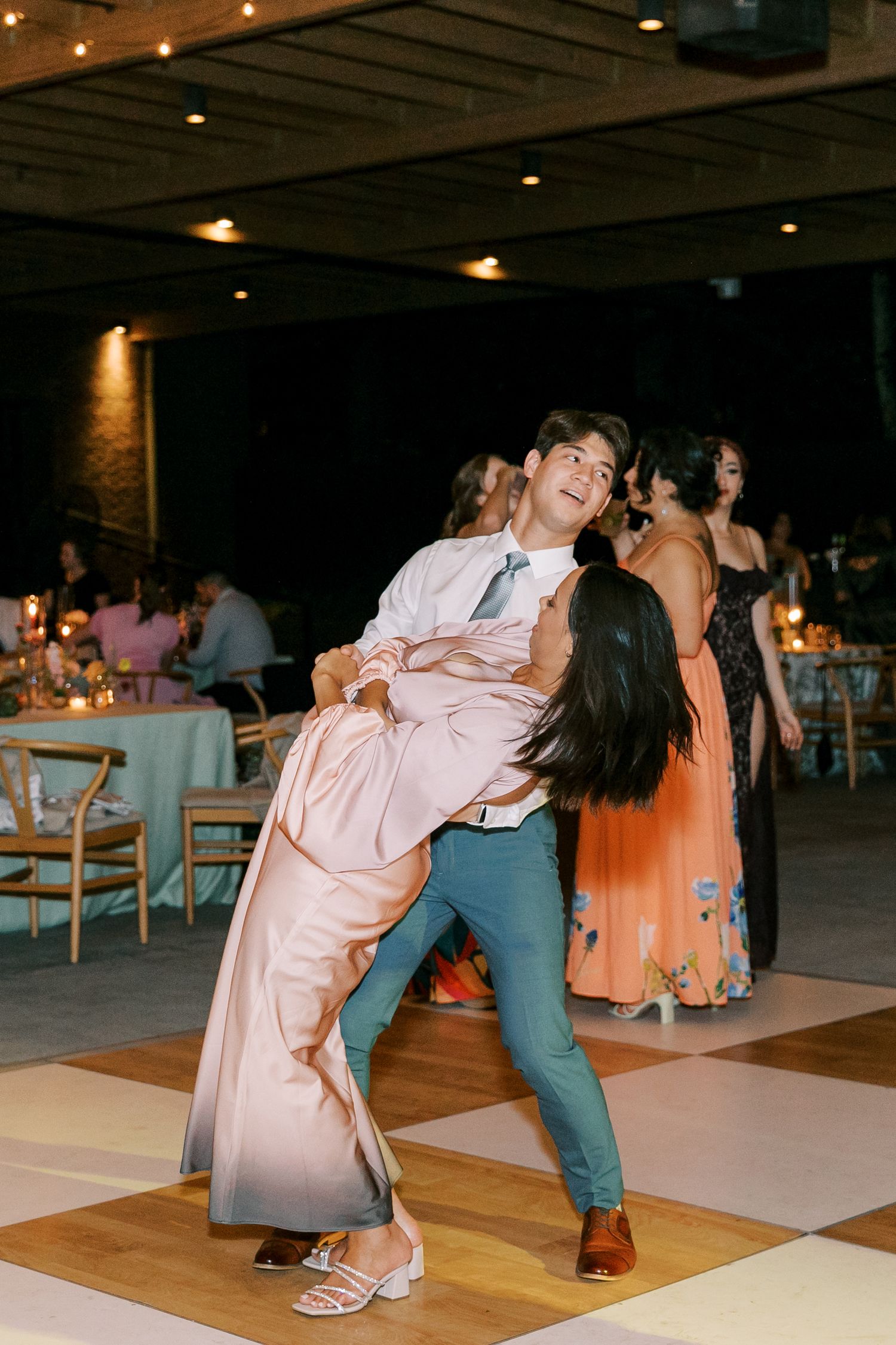 A couple performs an elegant dance move with a dramatic dip on a checkerboard dance floor at an indoor reception.