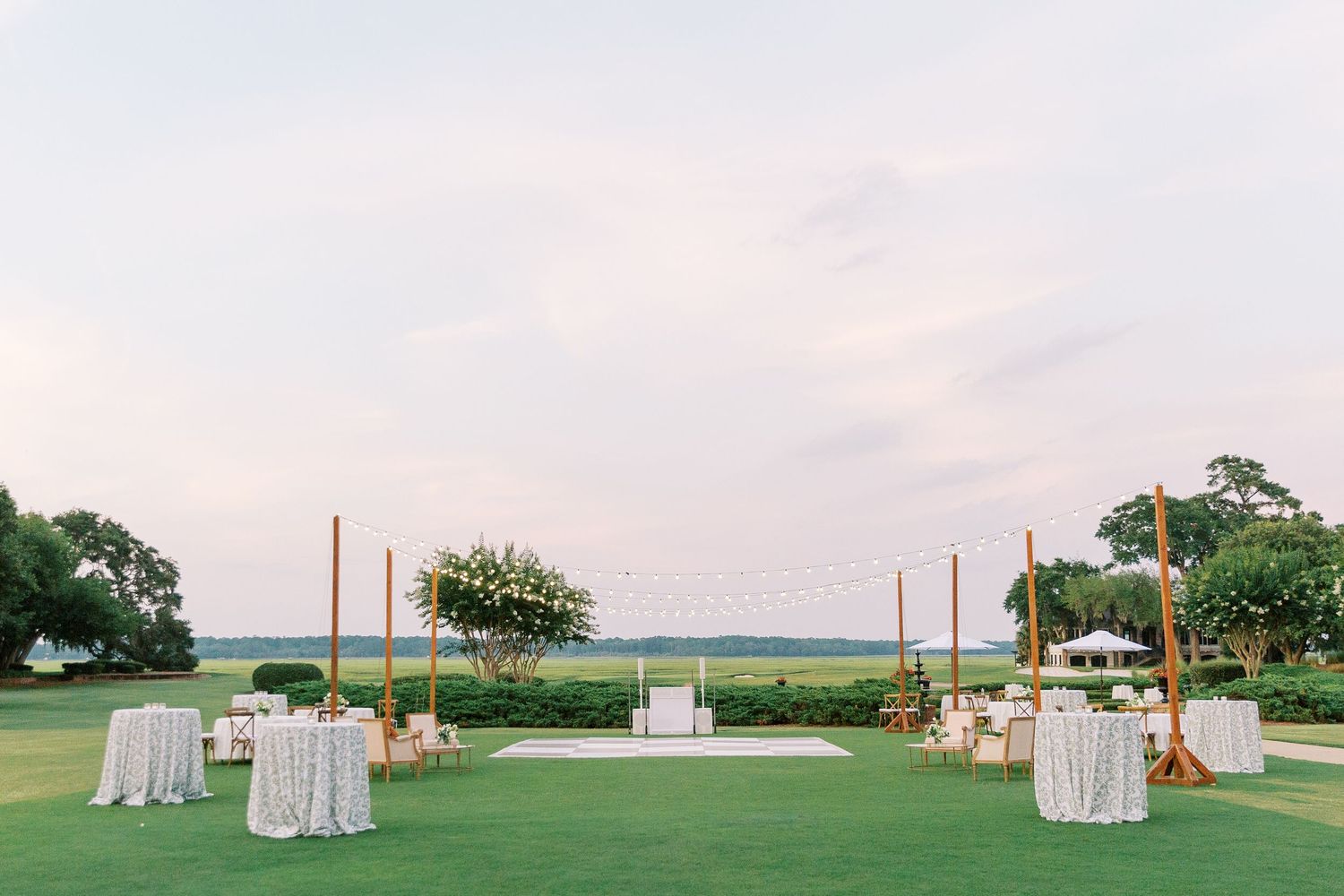 Outdoor wedding reception setup with white tables and wooden poles on grassy lawn at sunset.