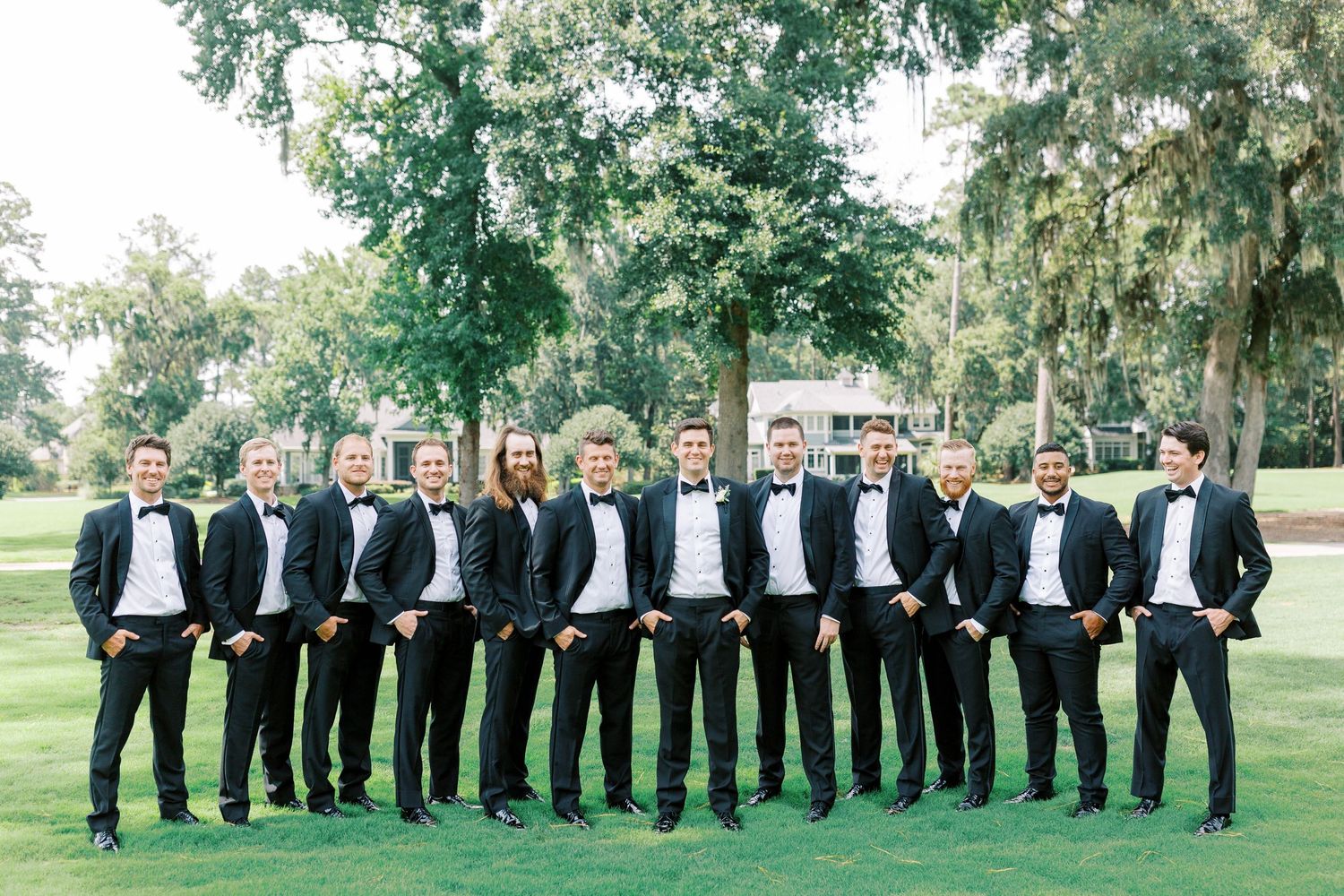 Group of groomsmen in matching black tuxedos and bow ties pose together on green lawn with trees in background.