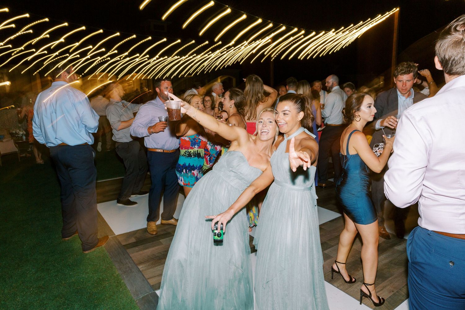 Wedding guests in blue dresses twirl and dance under string lights at an outdoor reception.
