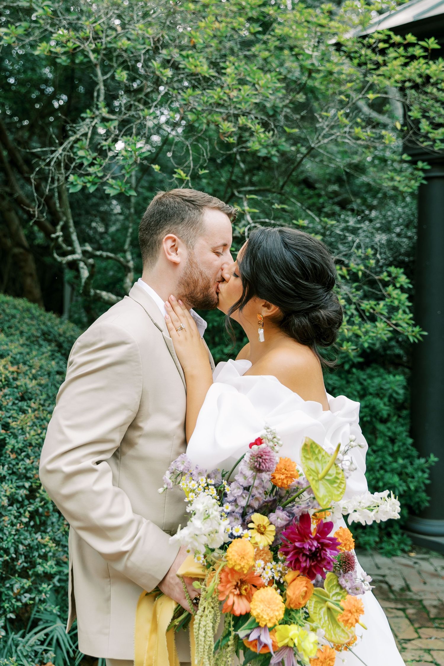 A tender wedding kiss moment captured in a lush garden setting with a colorful wildflower bouquet.