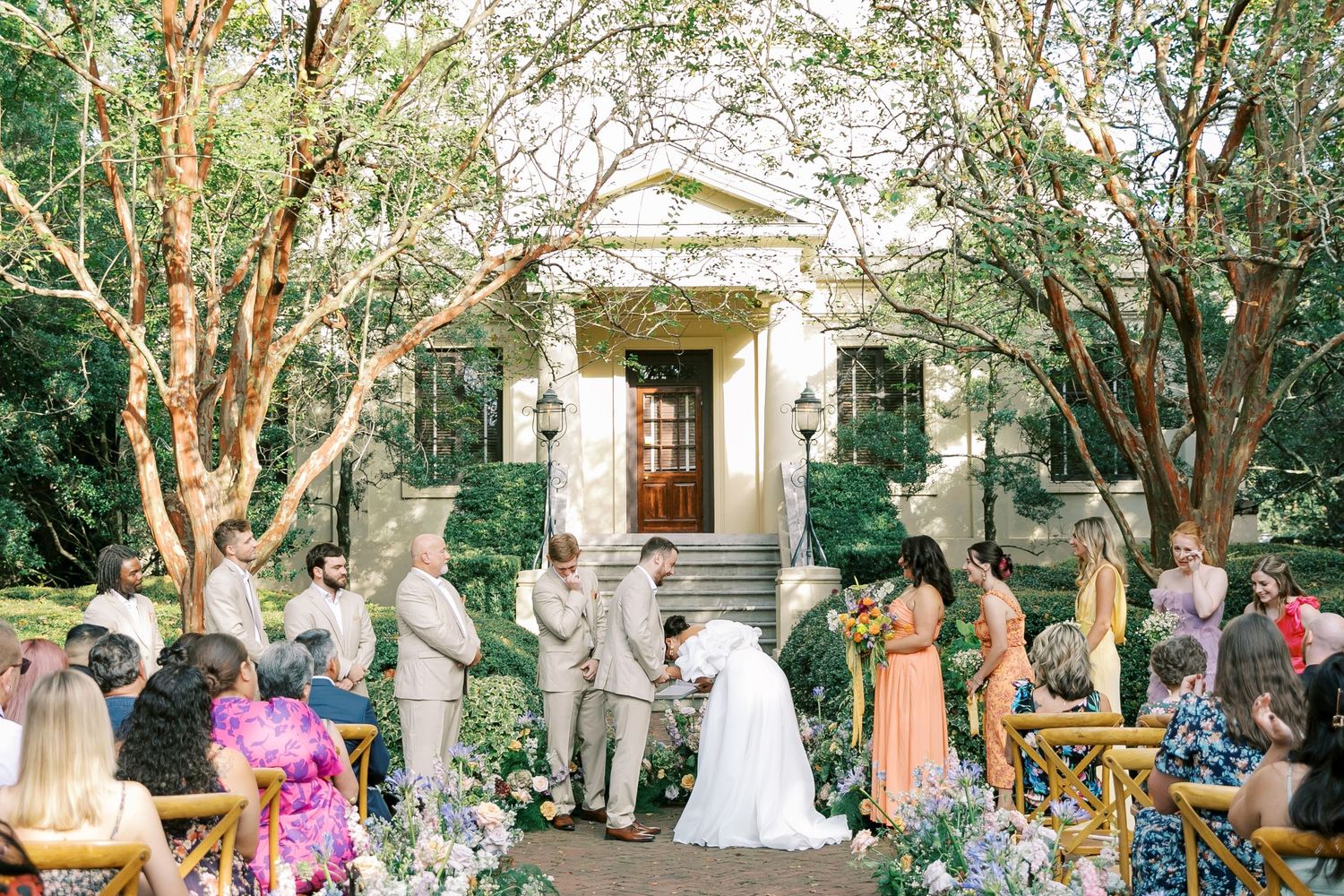 Wedding ceremony group gathers under lush green trees outside a historic stone building.