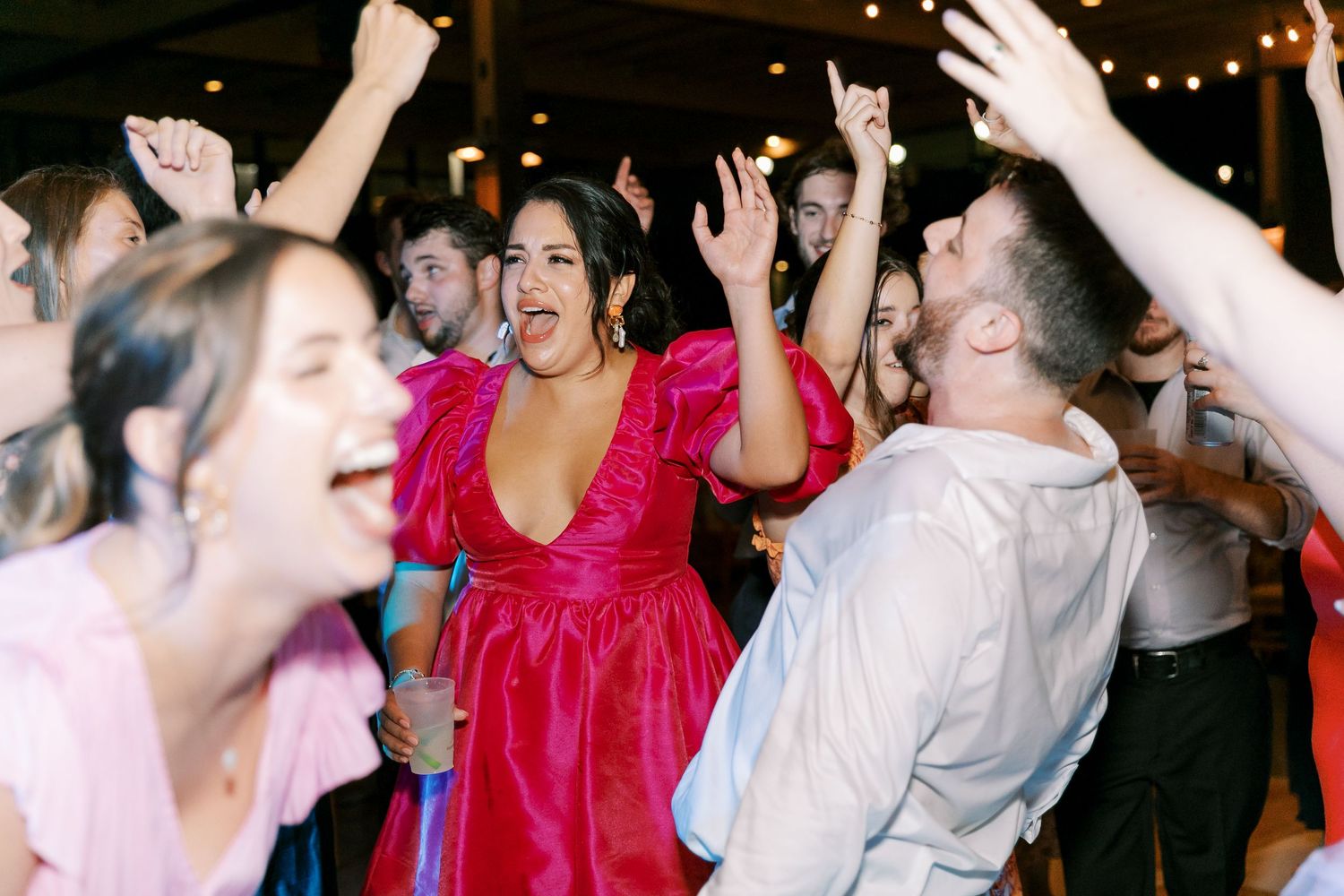 Joyful wedding guests dancing enthusiastically under string lights at an indoor reception.