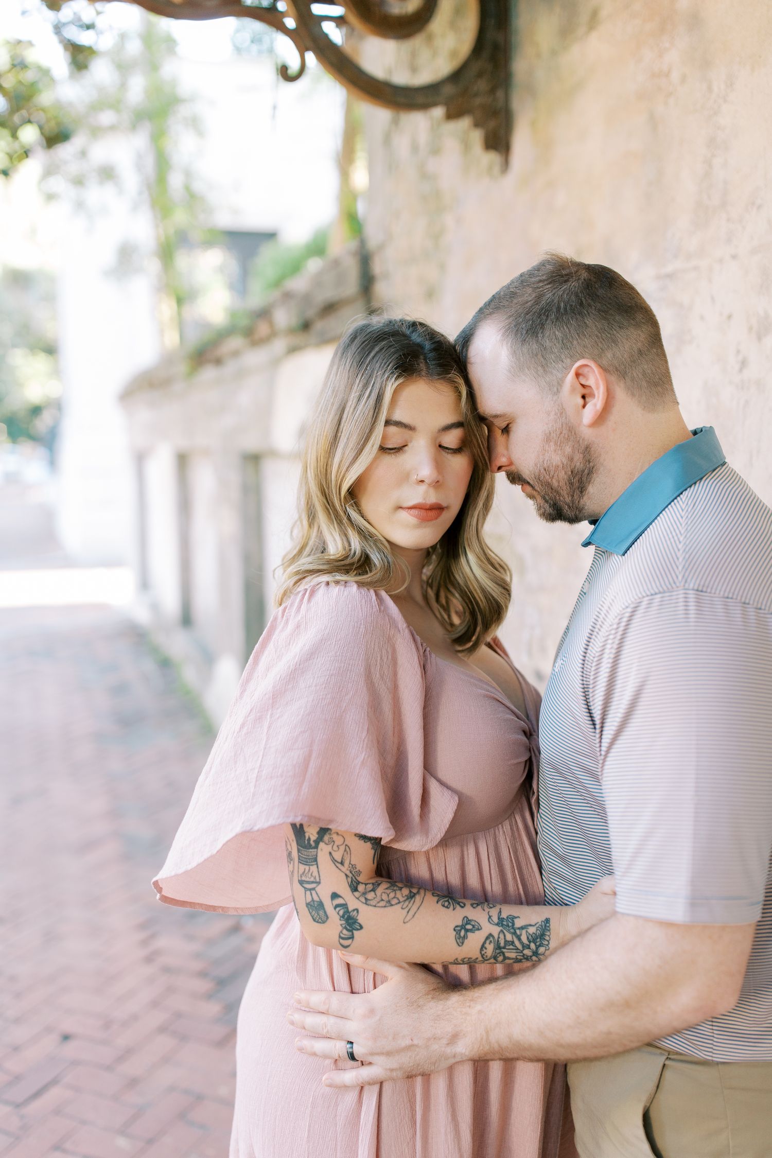 Romantic maternity photoshoot under rustic architecture with couple in pink and neutral tones.