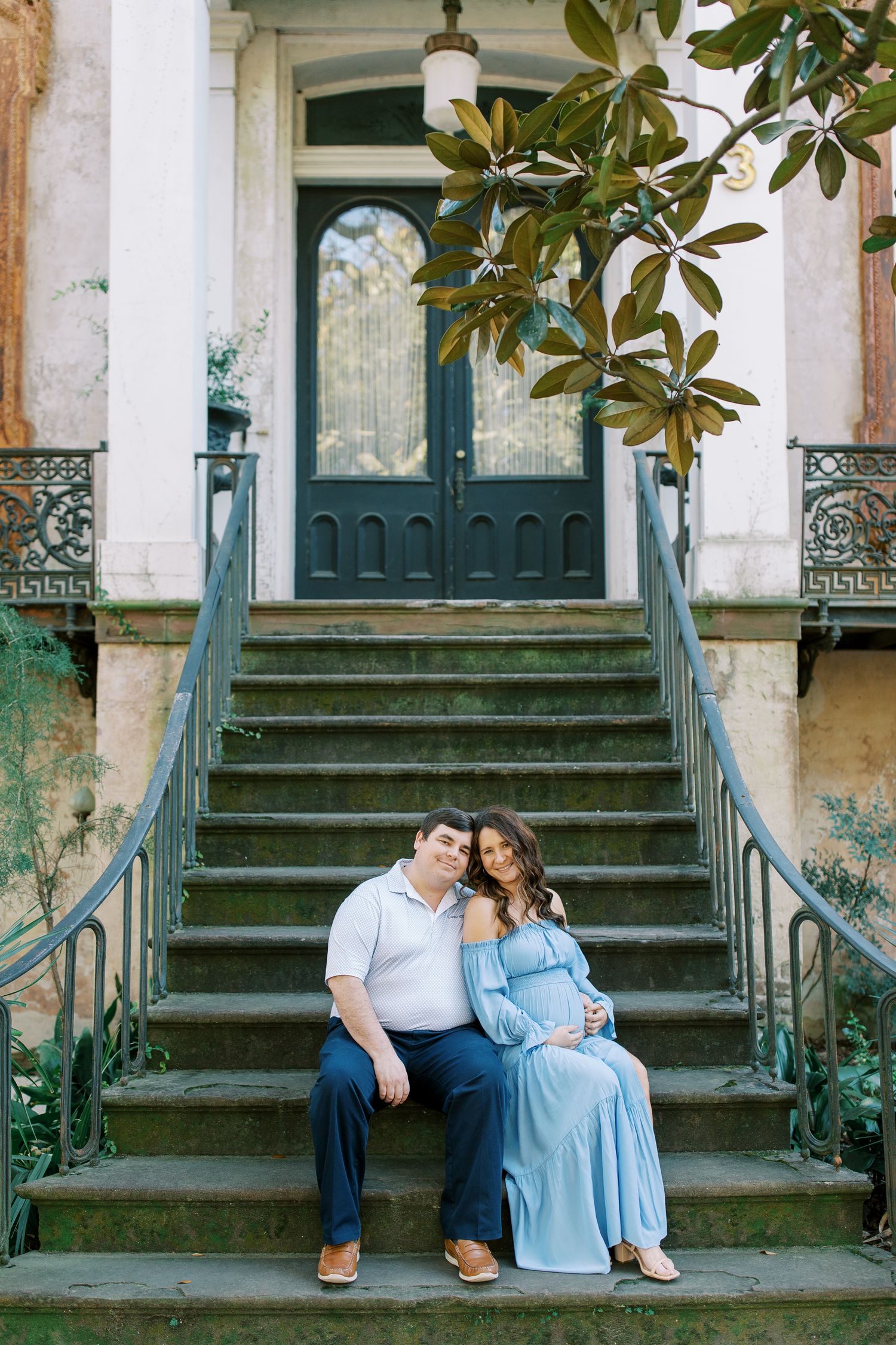 An expectant couple sits on historic mansion steps with ornate ironwork and magnolia leaves.