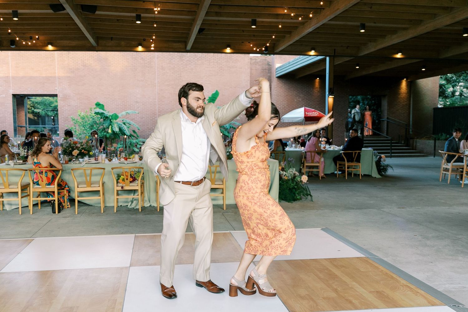 Couple dances joyfully on a dance floor at an indoor reception venue with exposed ceiling.