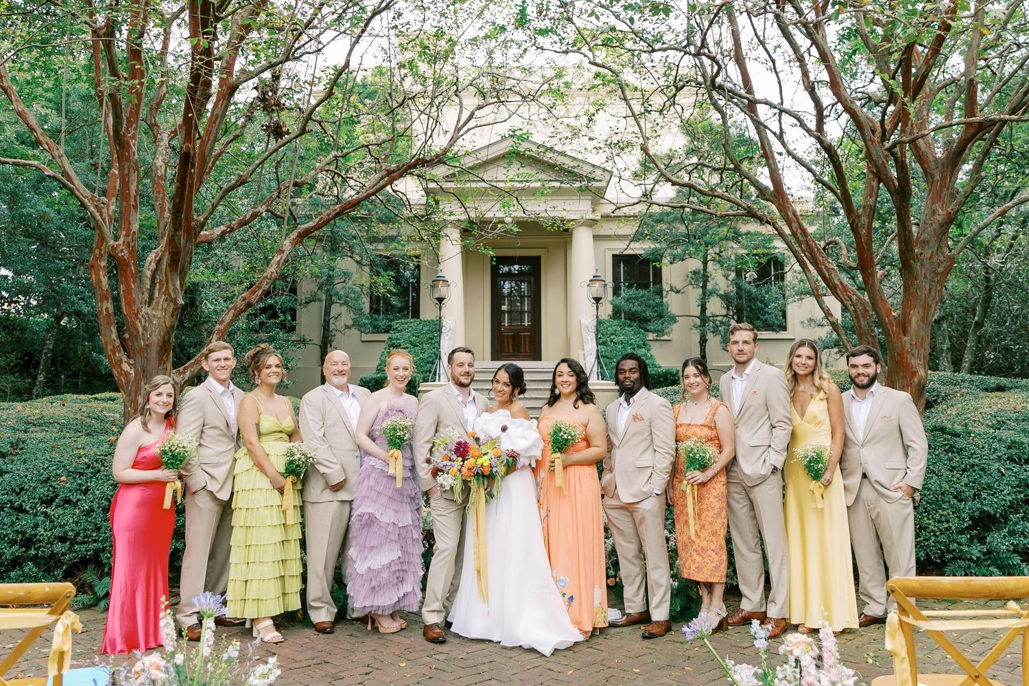 Wedding party wearing pastel colored dresses and suits poses together in front of a grand estate entrance.