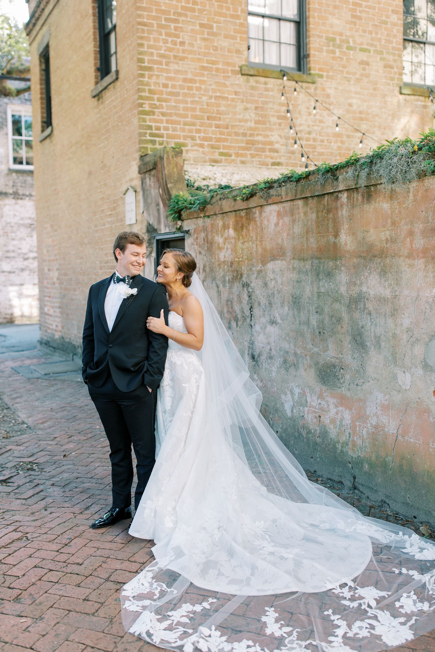 A romantic wedding couple shares tender moments on a brick pathway next to a stone wall.