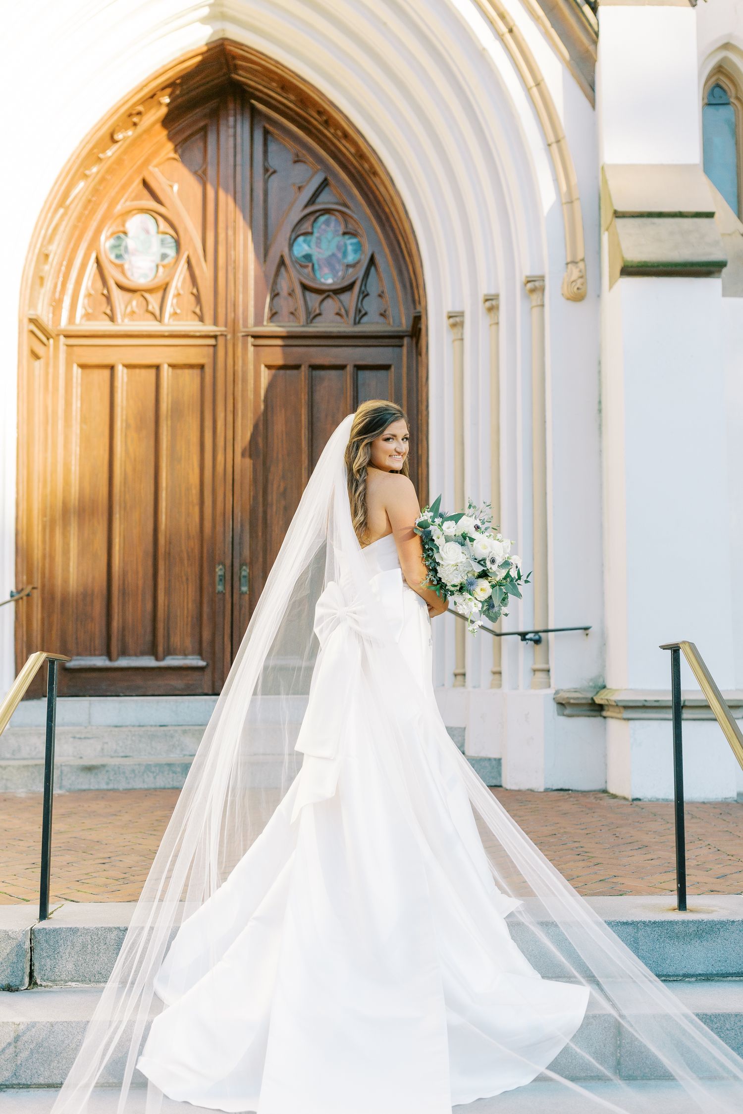 A bride in a flowing white wedding gown poses elegantly in front of ornate cathedral doors at a church entrance.