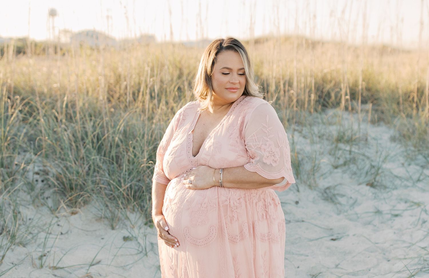 Person in flowy pink dress stands on sandy beach dunes with tall grass and warm sunset lighting in background.
