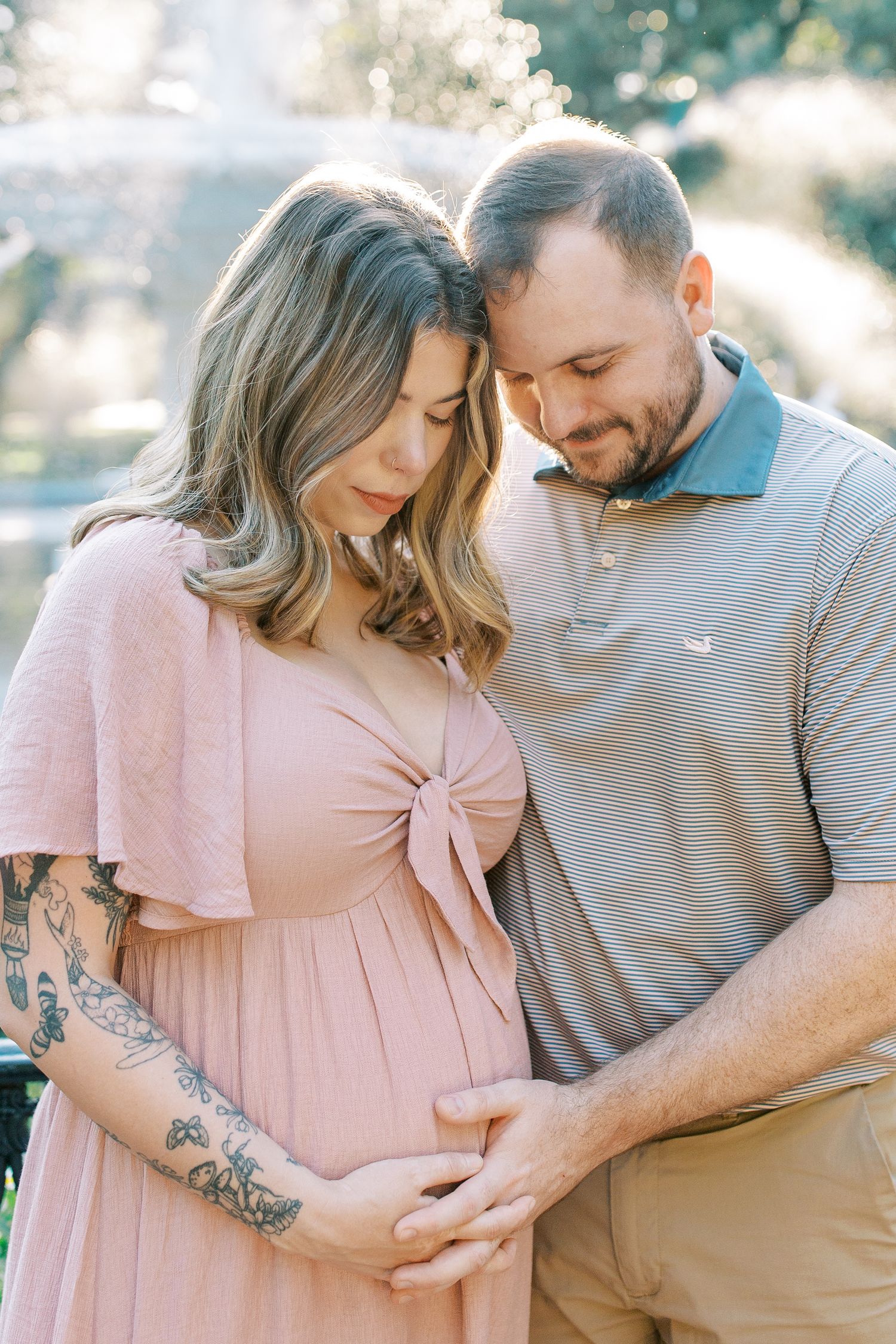 Expectant couple in pink dress and gray polo share tender moment outdoors while holding baby bump in sunlit setting in forsyth park