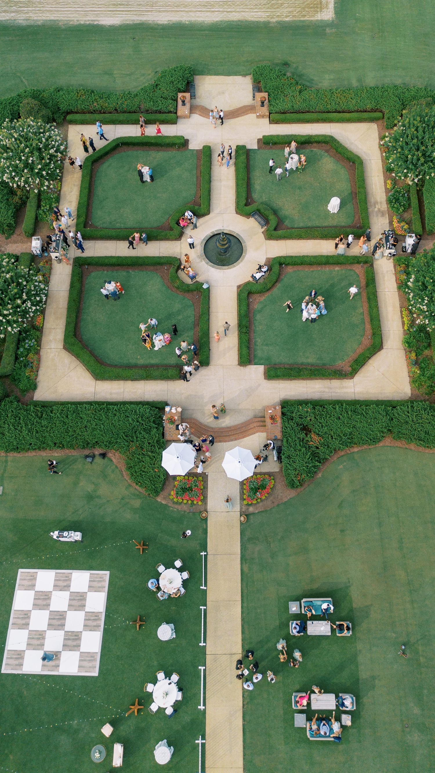 Aerial view of a symmetrical formal garden with neatly manicured lawns, pathways, and groups gathered for outdoor activities.
