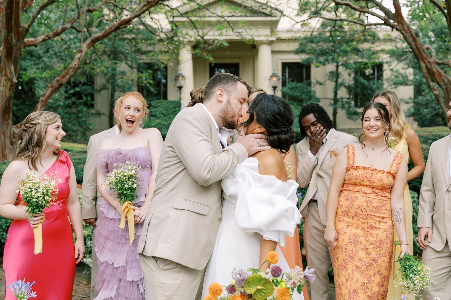 Wedding party poses outdoors in front of columned building with bridesmaids in colorful dresses.