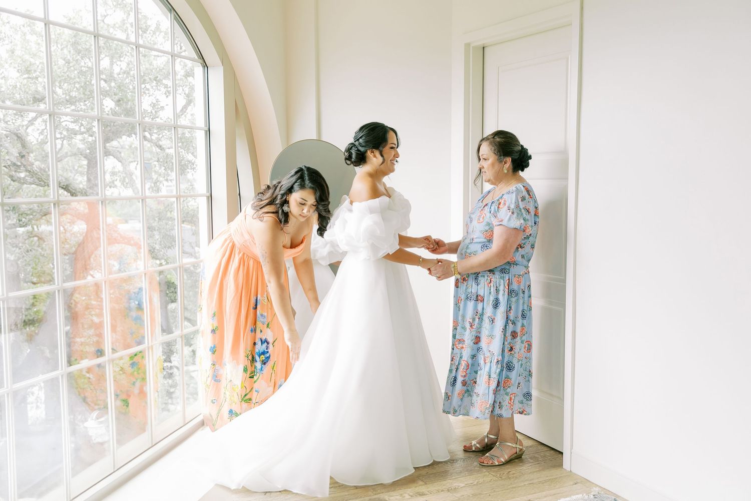 Bridesmaids in floral dresses help the bride get ready in a bright sunlit room.