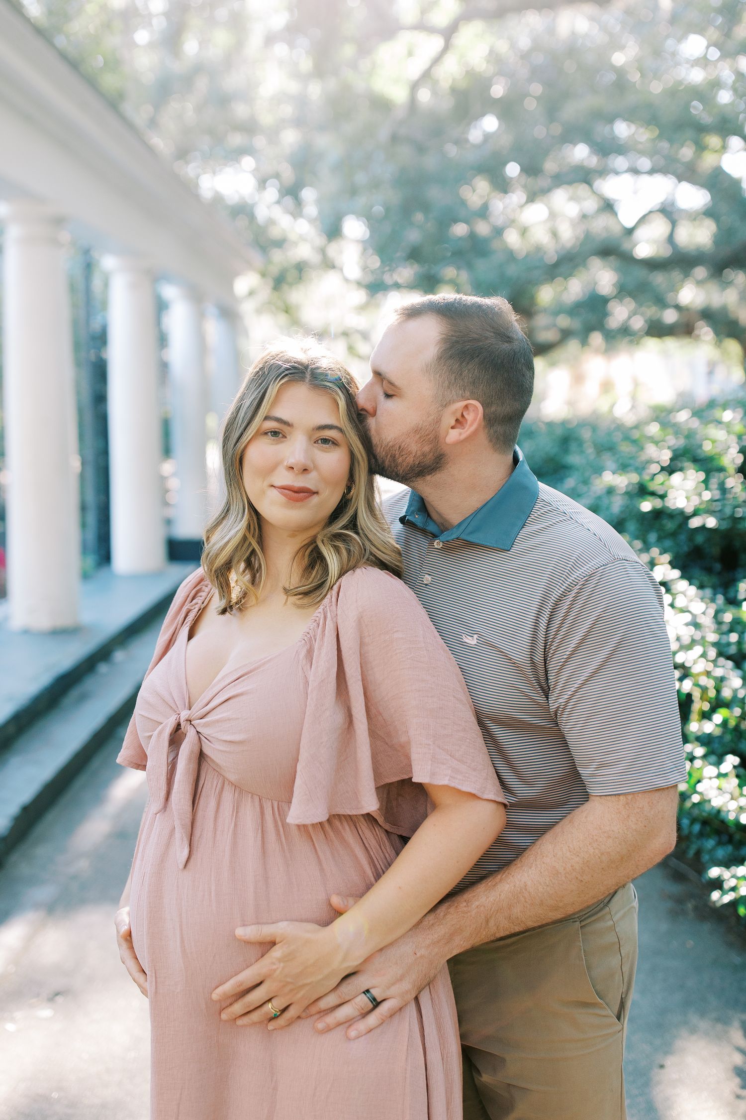 A tender maternity photo featuring a couple posing together on a porch with white columns in soft natural lighting.