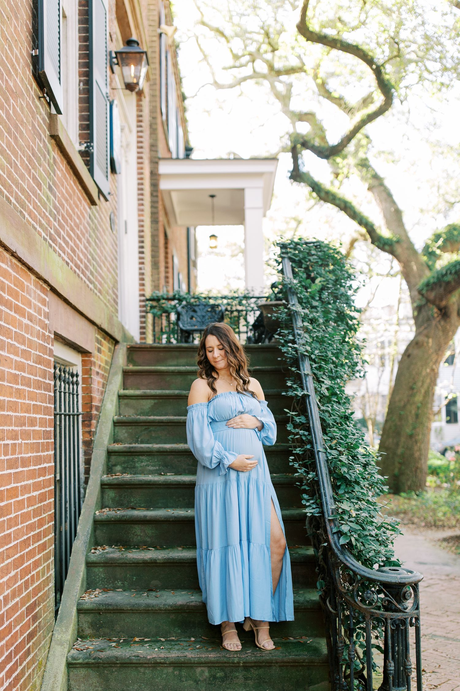A woman in a flowing blue dress stands on brick steps beside an elegant brick building with ivy.