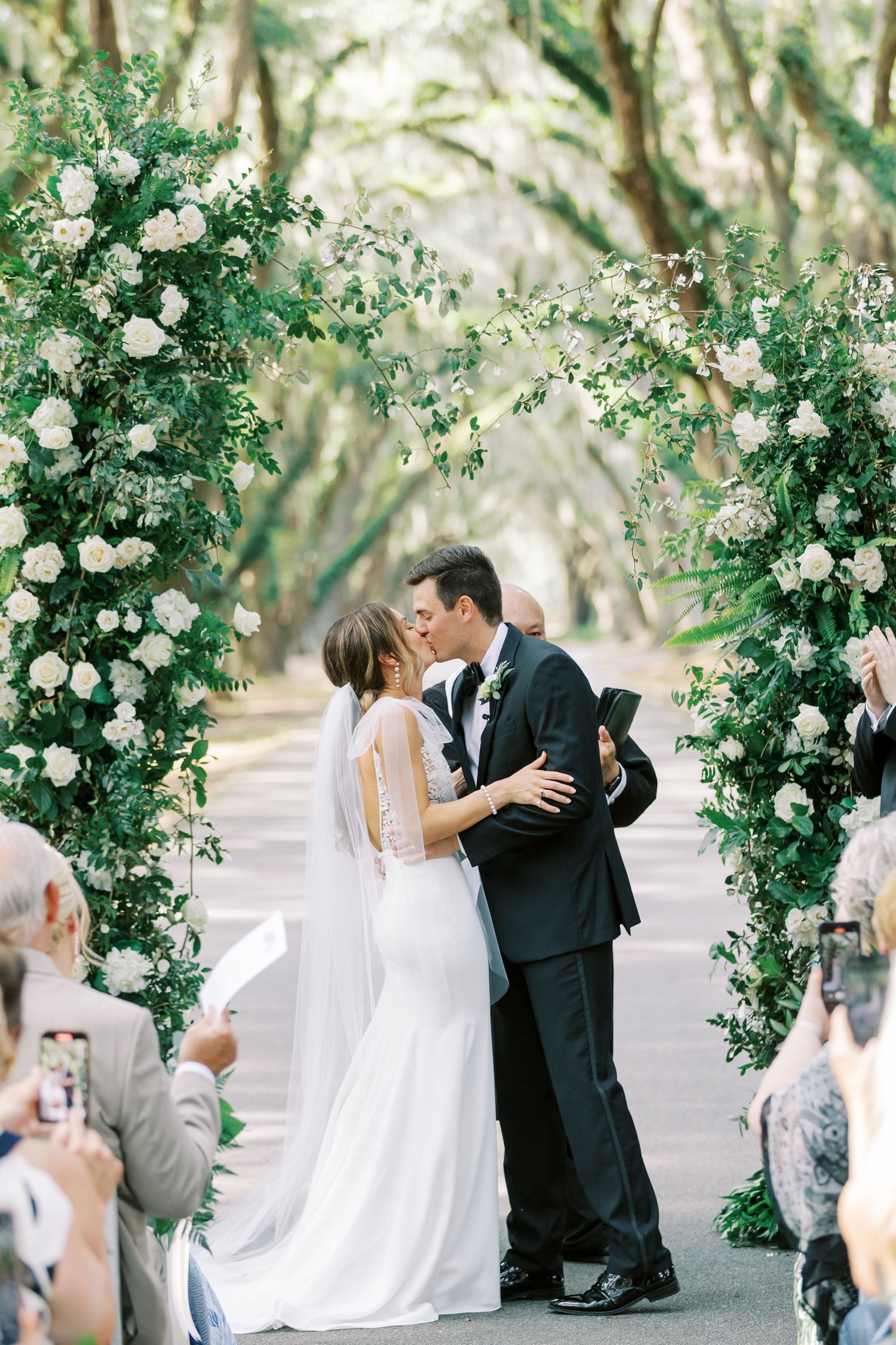 Couple sharing first kiss under floral arch during outdoor garden ceremony.