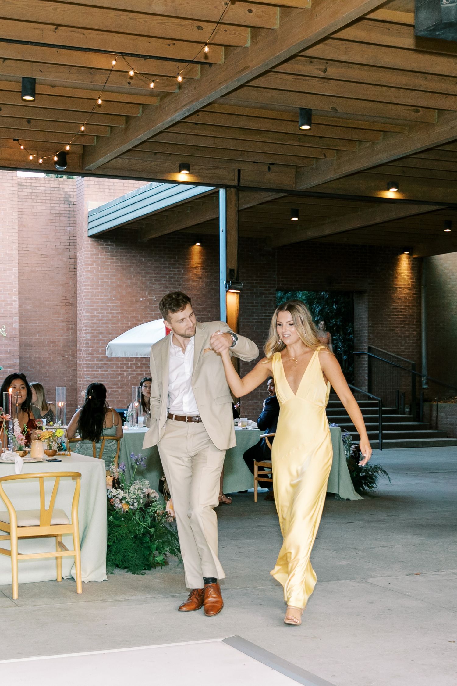 Wedding party members dance and celebrate in a covered outdoor venue with string lights.