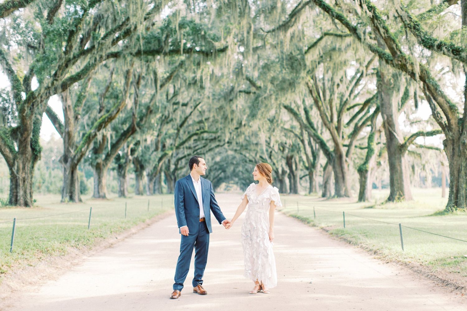Two people hold hands while walking down a romantic tree-lined path with Spanish moss hanging from old oaks at Wormsloe in Savannah.