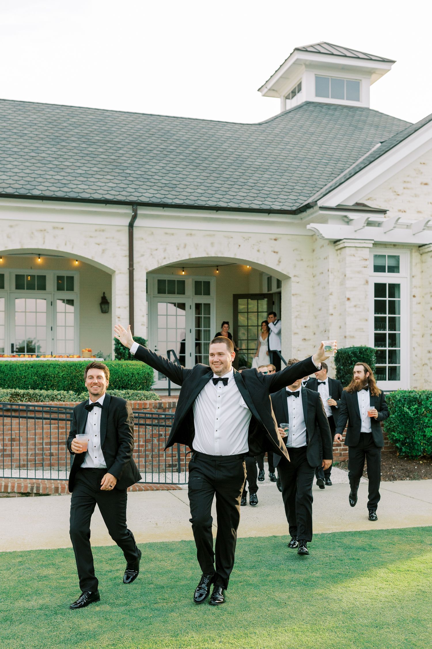 Groomsmen in tuxedos jump joyfully outside an elegant white building with arched doorways.