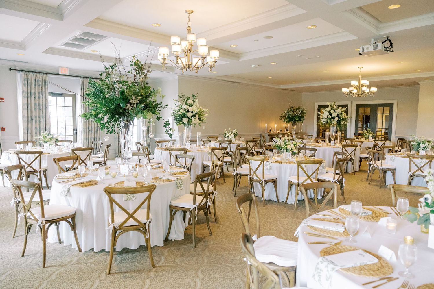 Elegant wedding reception venue with round tables, white linens, rustic wooden chairs and tall floral centerpieces.