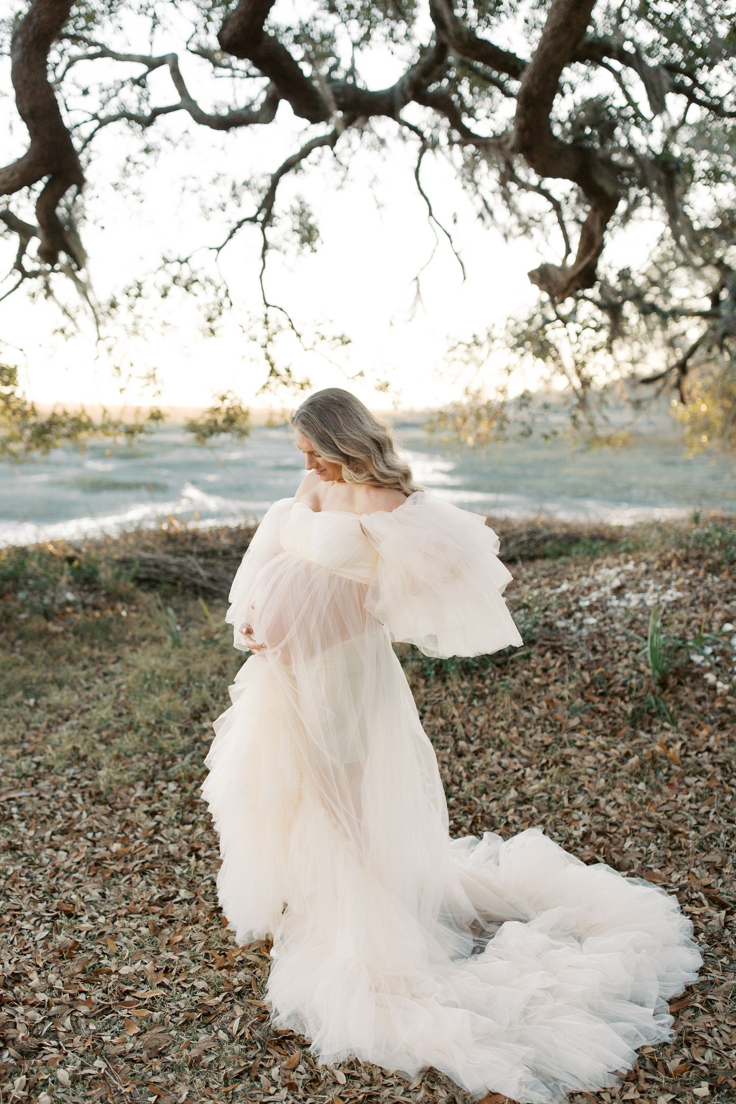 A romantic ethereal bridal portrait features a flowing off-white tulle gown beneath an oak tree at sunset.