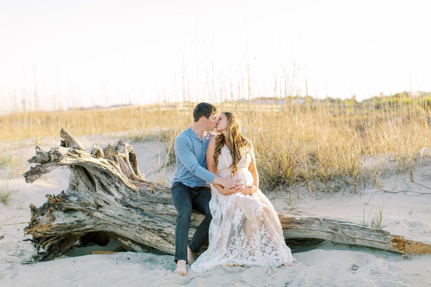 A romantic couple sits together on a large piece of driftwood at a beach with tall grass during sunset.