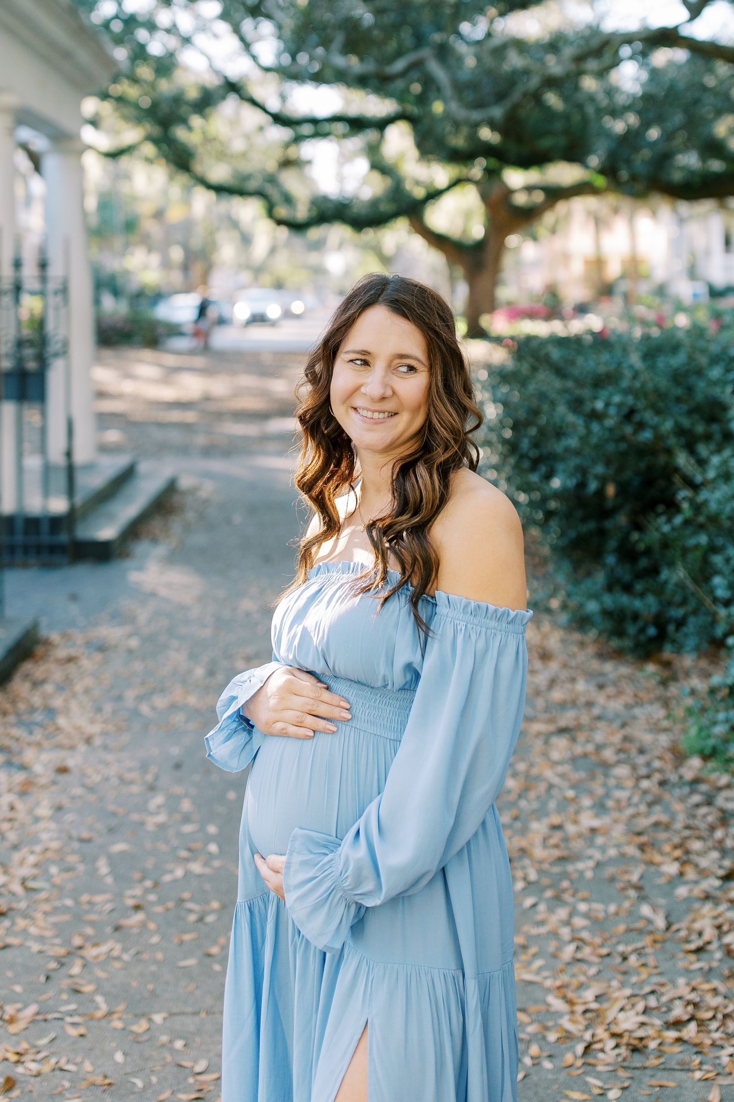A woman in a flowing light blue maternity dress poses on a tree-lined sidewalk.