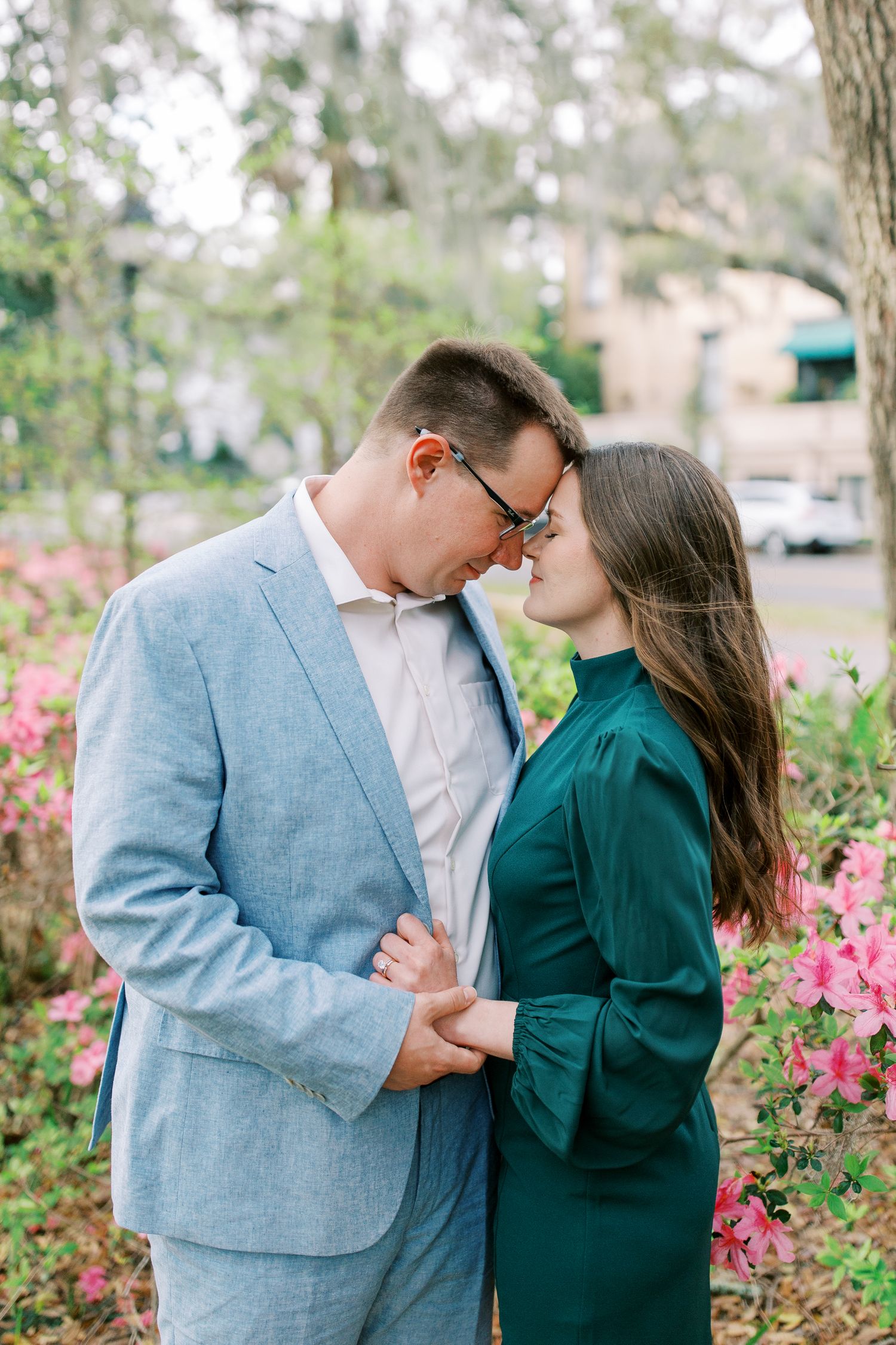Couple shares intimate moment near pink flowers with person in light blue suit and dark green dress.