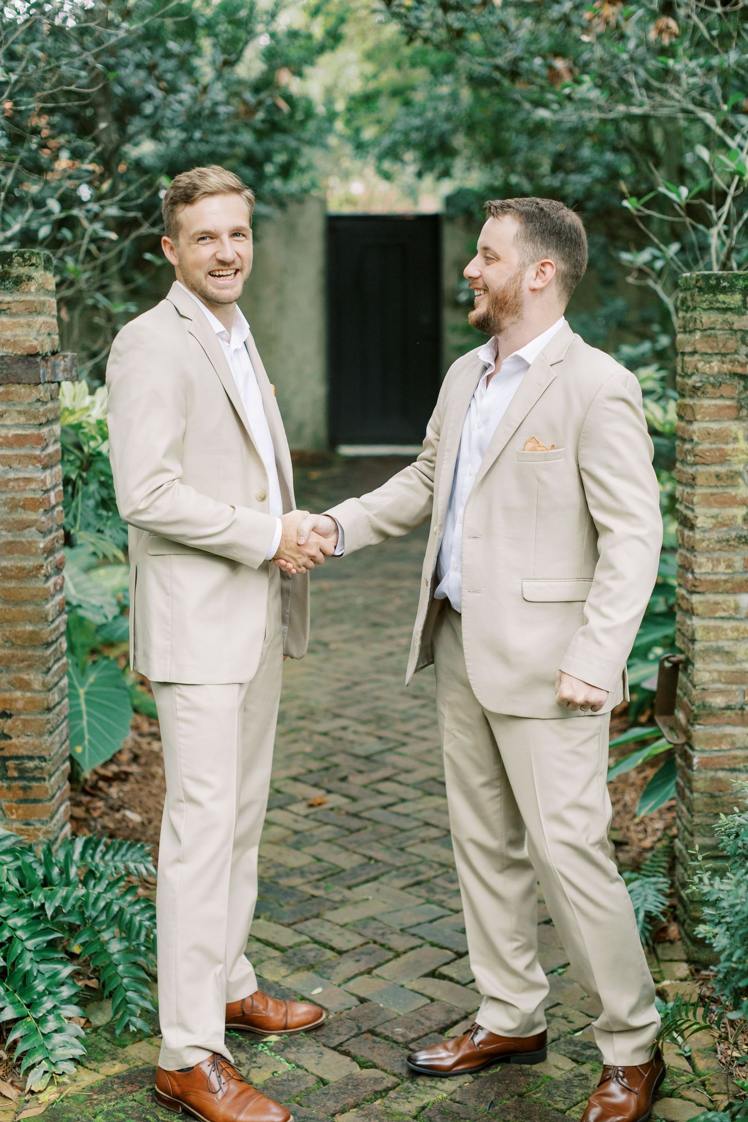 Two people in beige suits shake hands while smiling on a brick pathway surrounded by greenery.