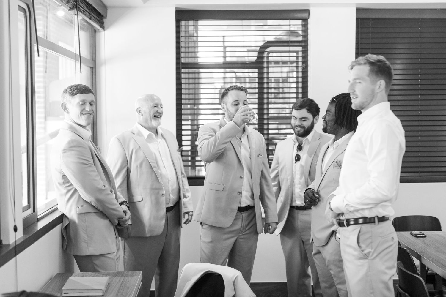 A group of people in business attire gather by a window in an office having a discussion.
