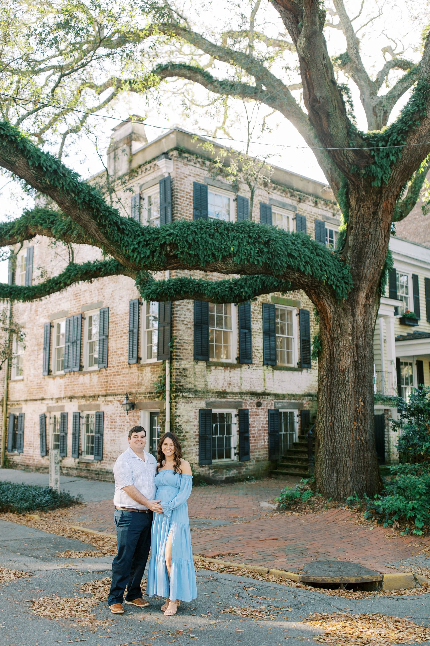 A couple poses in front of a historic building with large oak trees and brick sidewalks.